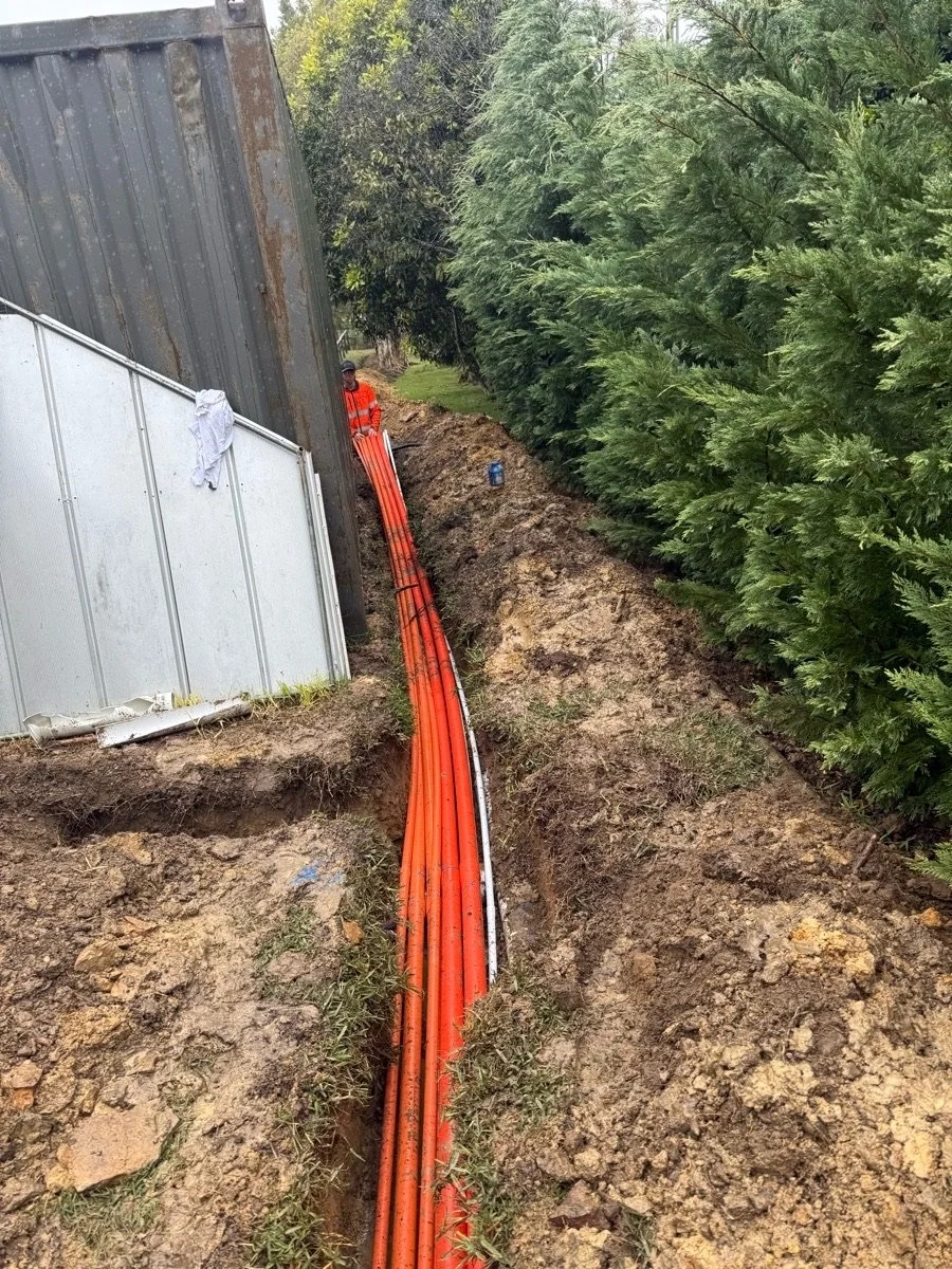 Construction worker in an orange safety uniform working on underground orange and white pipes along a trench surrounded by dirt and green trees.