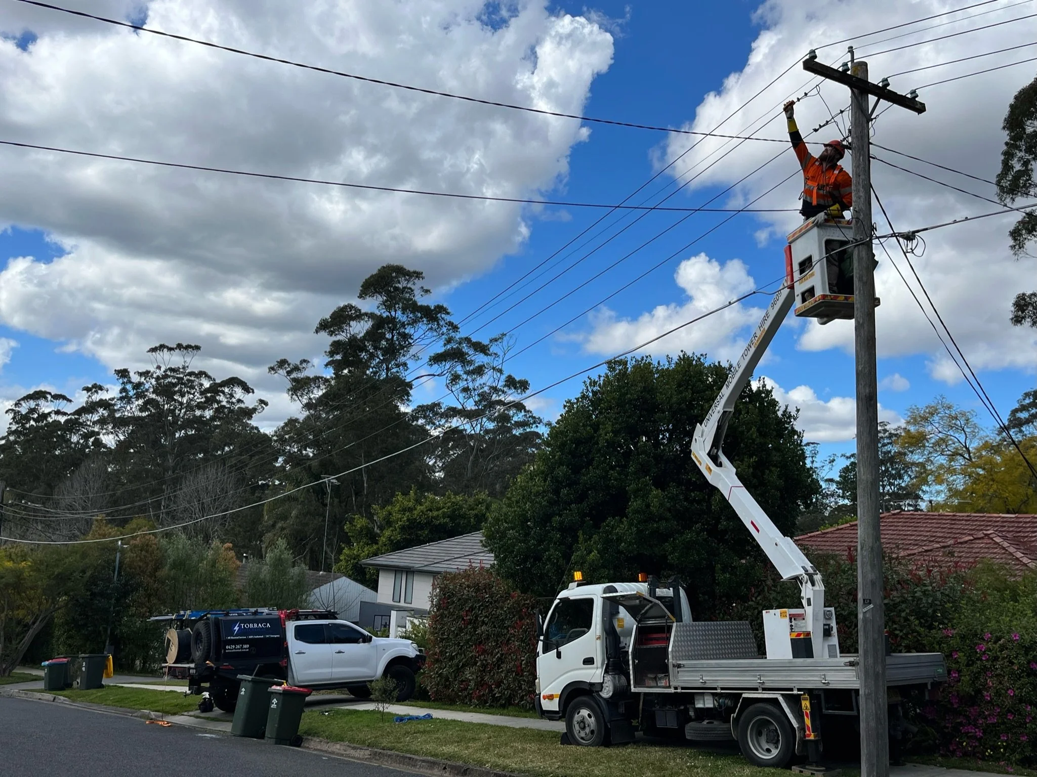 A utility worker in an orange vest is working on electric power lines while standing in an elevated bucket on a white utility truck. The truck is parked on a residential street lined with trees, houses, and a few parked vehicles, with a partly cloudy