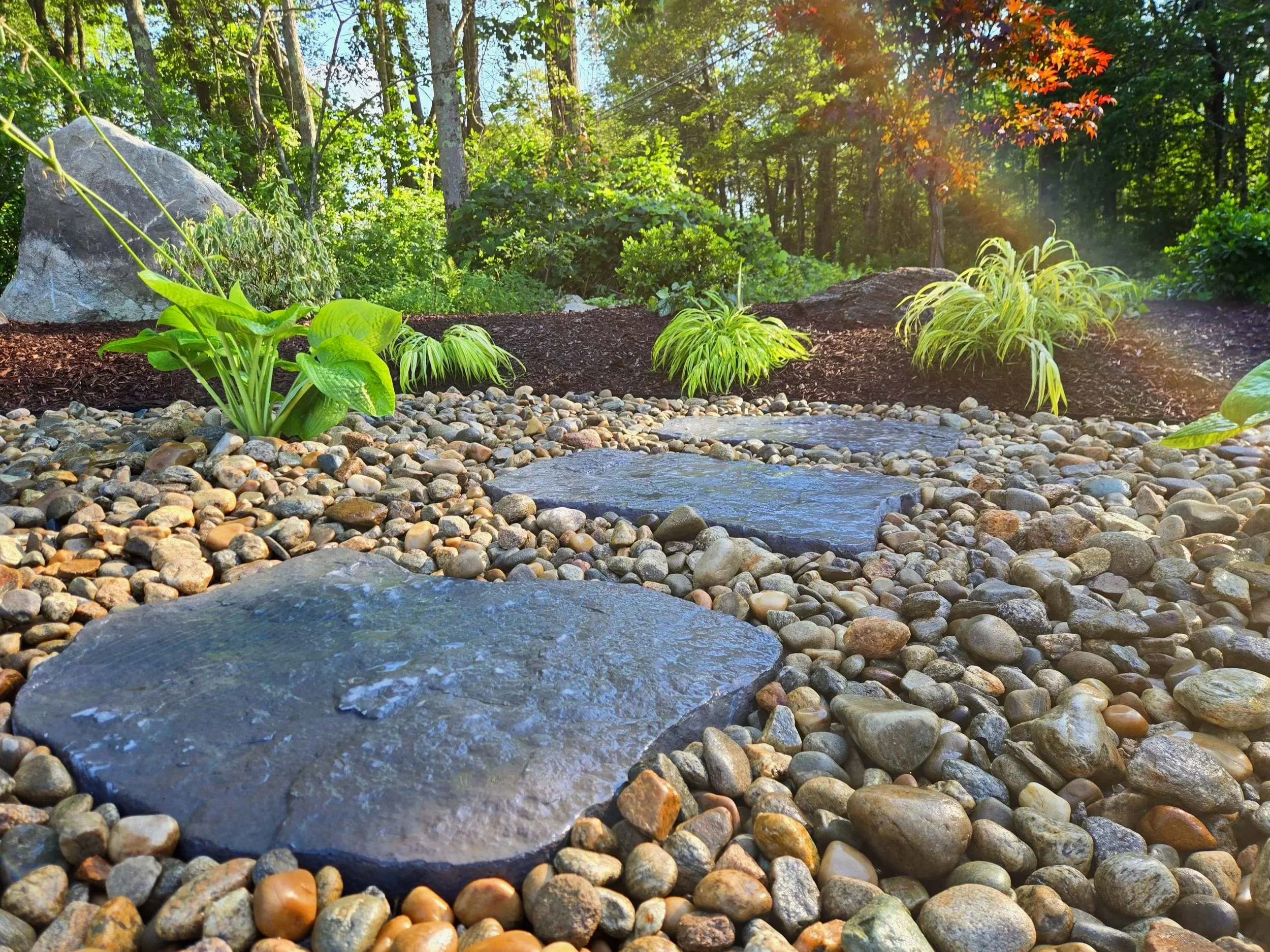 A landscaped garden pathway with large flat stones surrounded by small pebbles, green plants, mulch, and a forested background under sunlight.   In Danbury CT and surrounding CT/NY counties such as Fairfield, Hartford, New Haven, Westchester, Putnam.