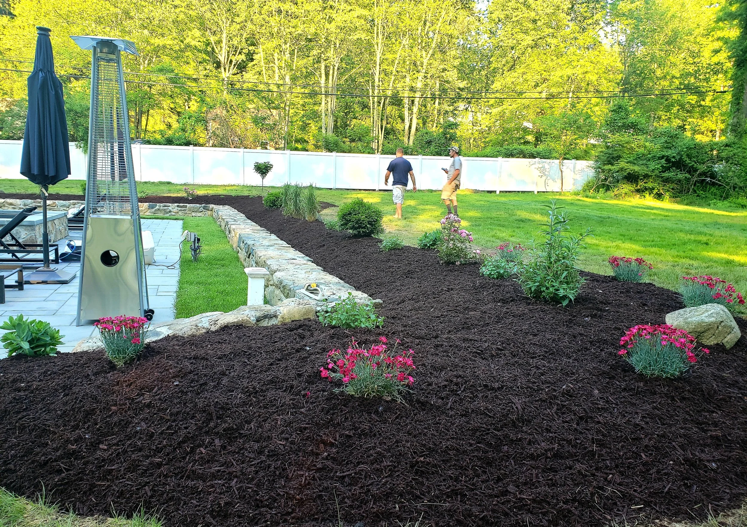 Two men gardening in a backyard with flowers and shrubs, with a stone border, a patio with a closed umbrella, and trees in the background.  Landscaping in Danbury CT and surrounding CT/NY counties such as Fairfield, Hartford, New Haven, Westchester,