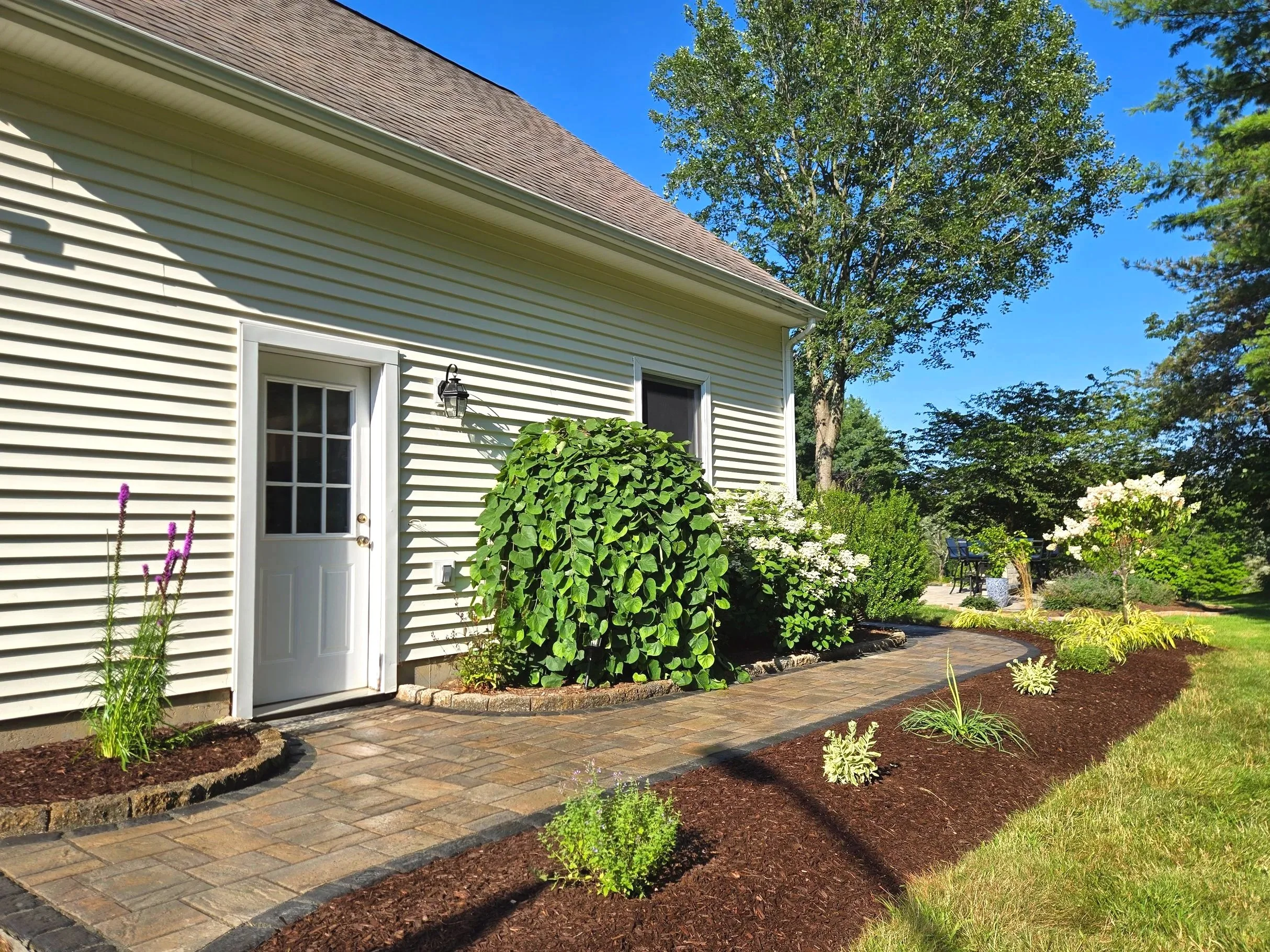 Backyard garden with a brick pathway, flowering bushes, and patio table and chairs.   Landscape design, installation, and consultation in Danbury CT and surrounding CT/NY counties such as Fairfield, Hartford, New Haven, Westchester, Putnam.