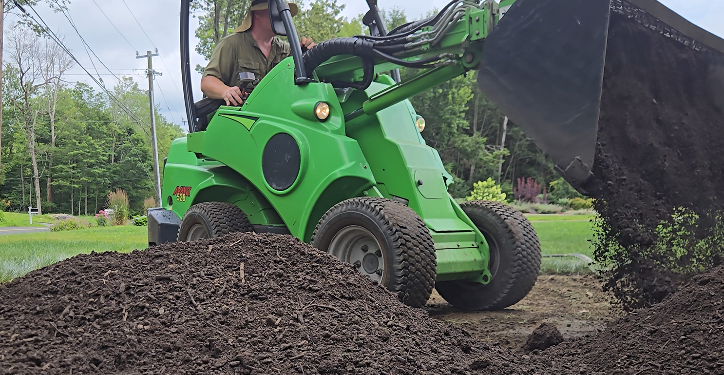 A person operating a bright green compact loader machine moving dirt outdoors during daytime, with trees and power lines in the background.   In Danbury CT and surrounding CT/NY counties such as Fairfield, Hartford, New Haven, Westchester, Putnam.