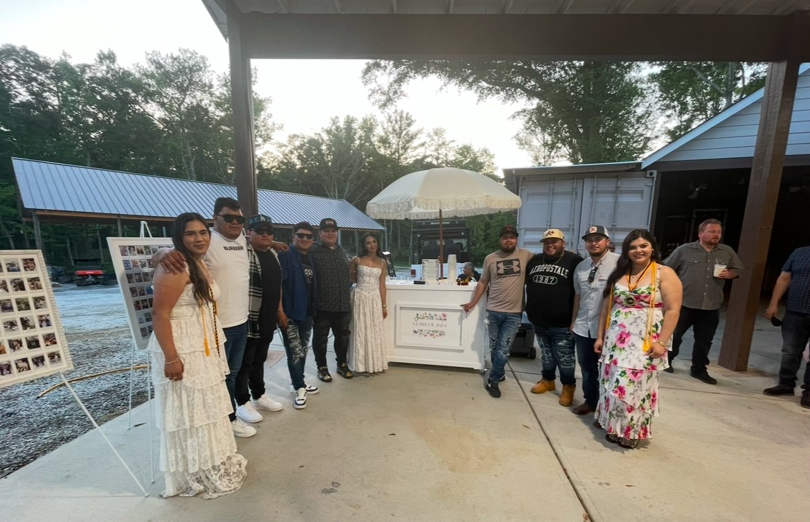Group of people posing outdoors in front of a white booth with a large parasol, wearing casual and semi-formal attire, with photo boards on easels nearby.