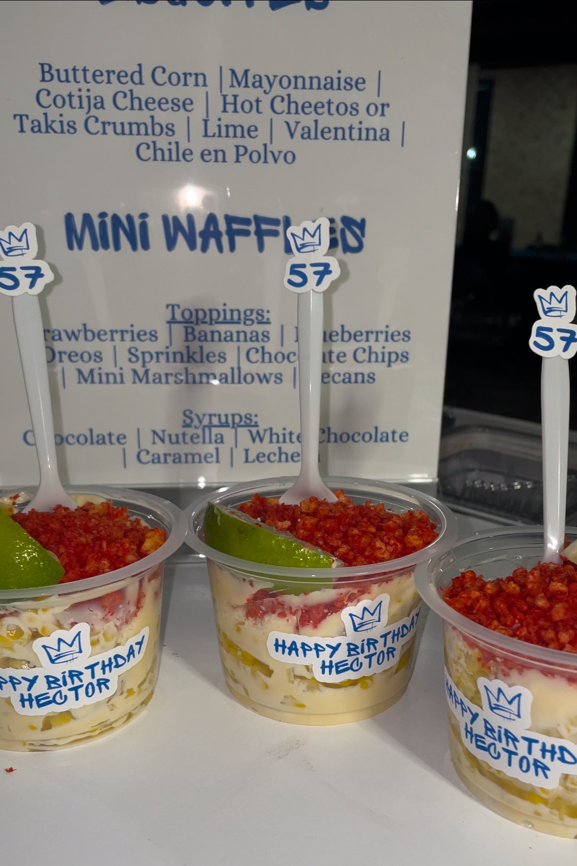 Cups of Mexican street corn with toppings, including lime and spicy crumbs, labeled for a birthday event.
