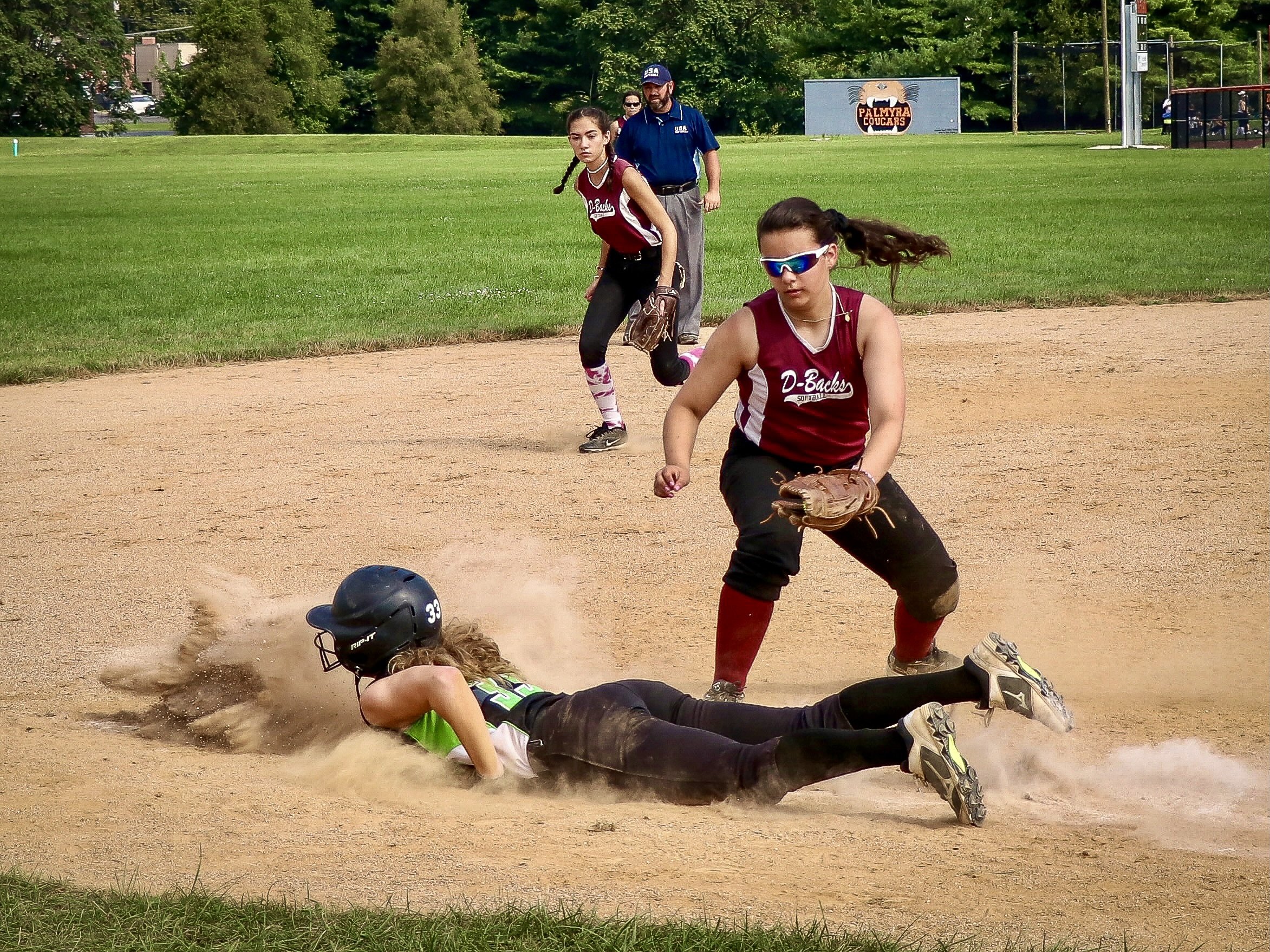 Softball runner in a black helmet slides into base through a cloud of dust as a fielder in sunglasses crouches with her glove ready, an infielder and umpire watching behind them