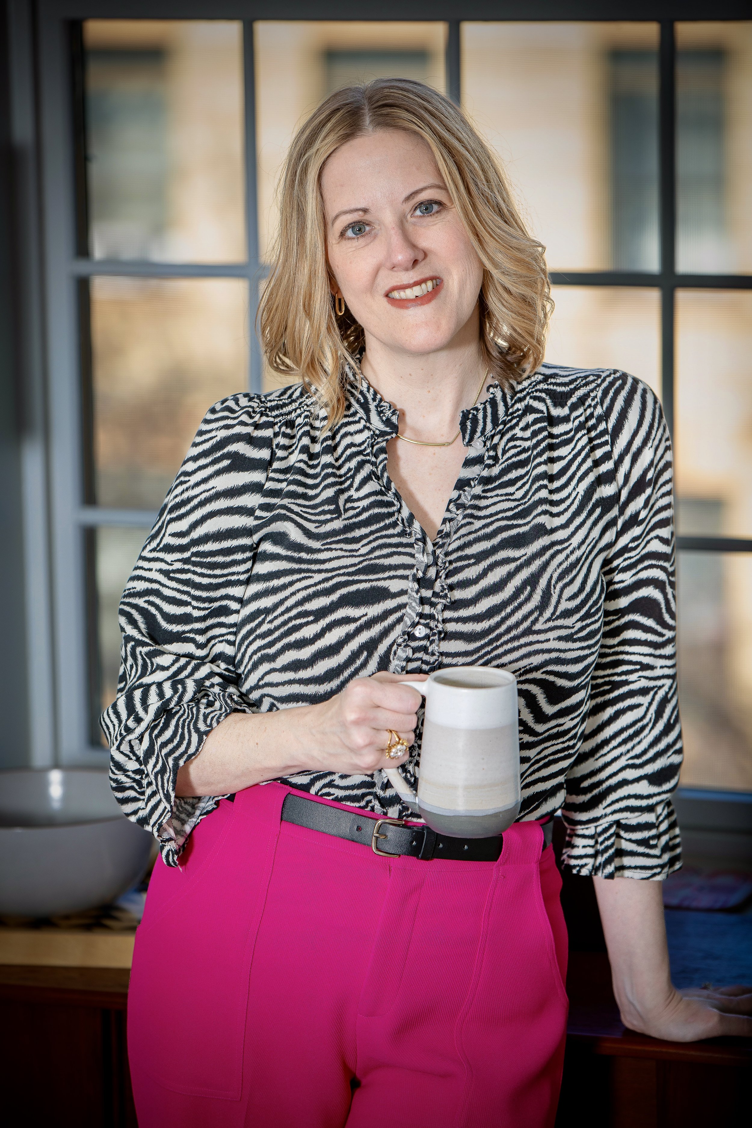 Woman smiling in a relaxed personal branding portrait, holding a coffee mug in natural window light