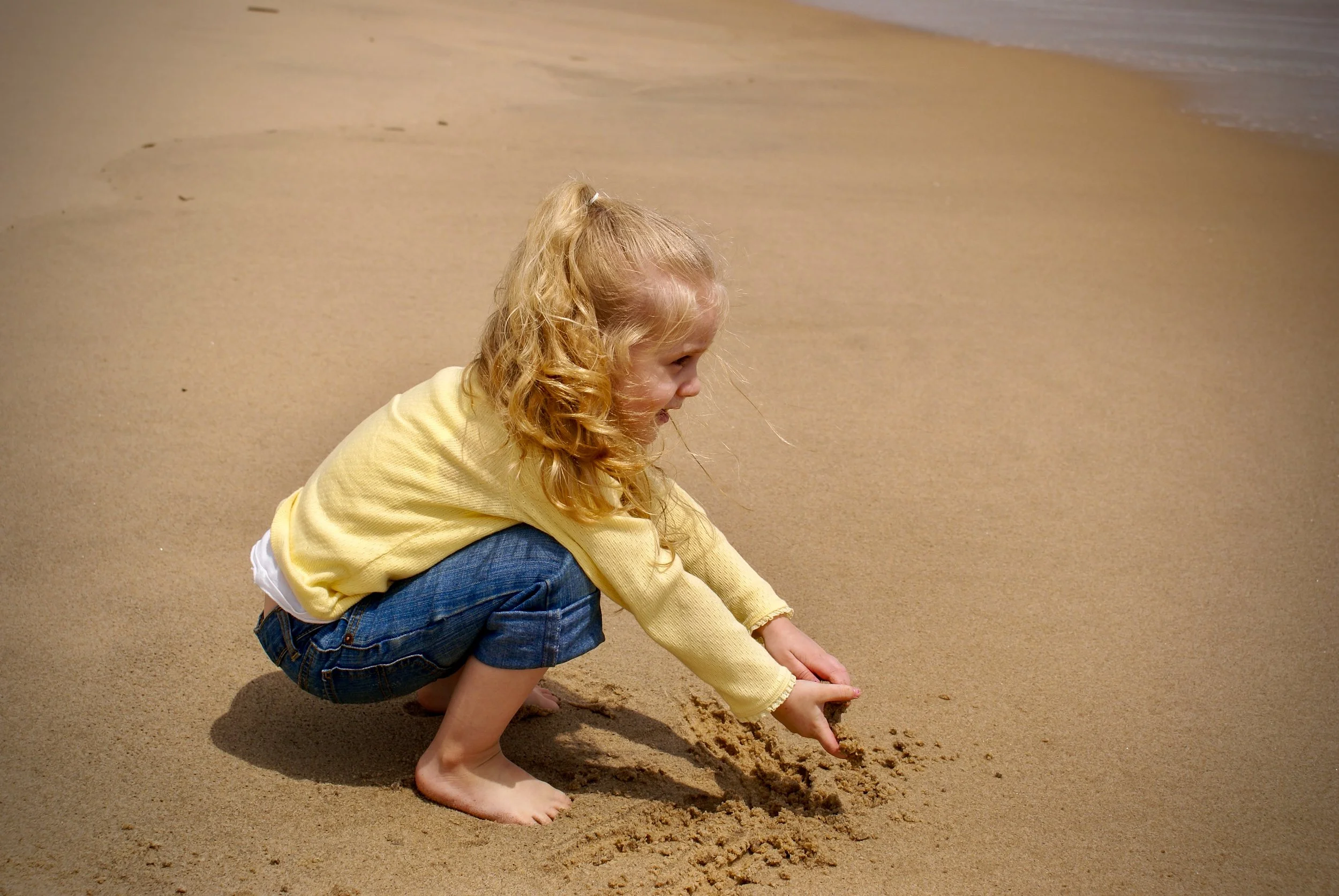 Toddler in a yellow sweater crouched barefoot on the beach, smiling as she digs in the sand