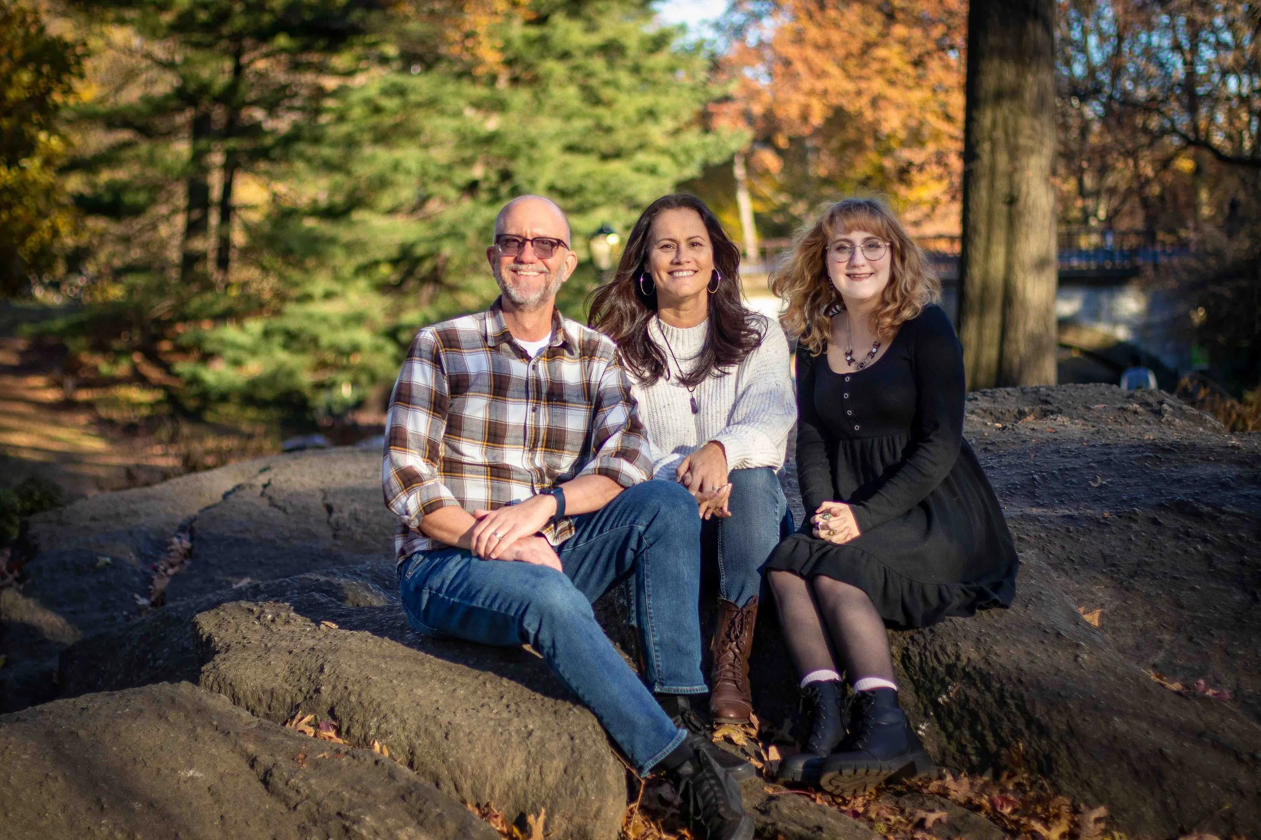 Family of three seated together on large rocks in a park, smiling, with fall foliage behind them