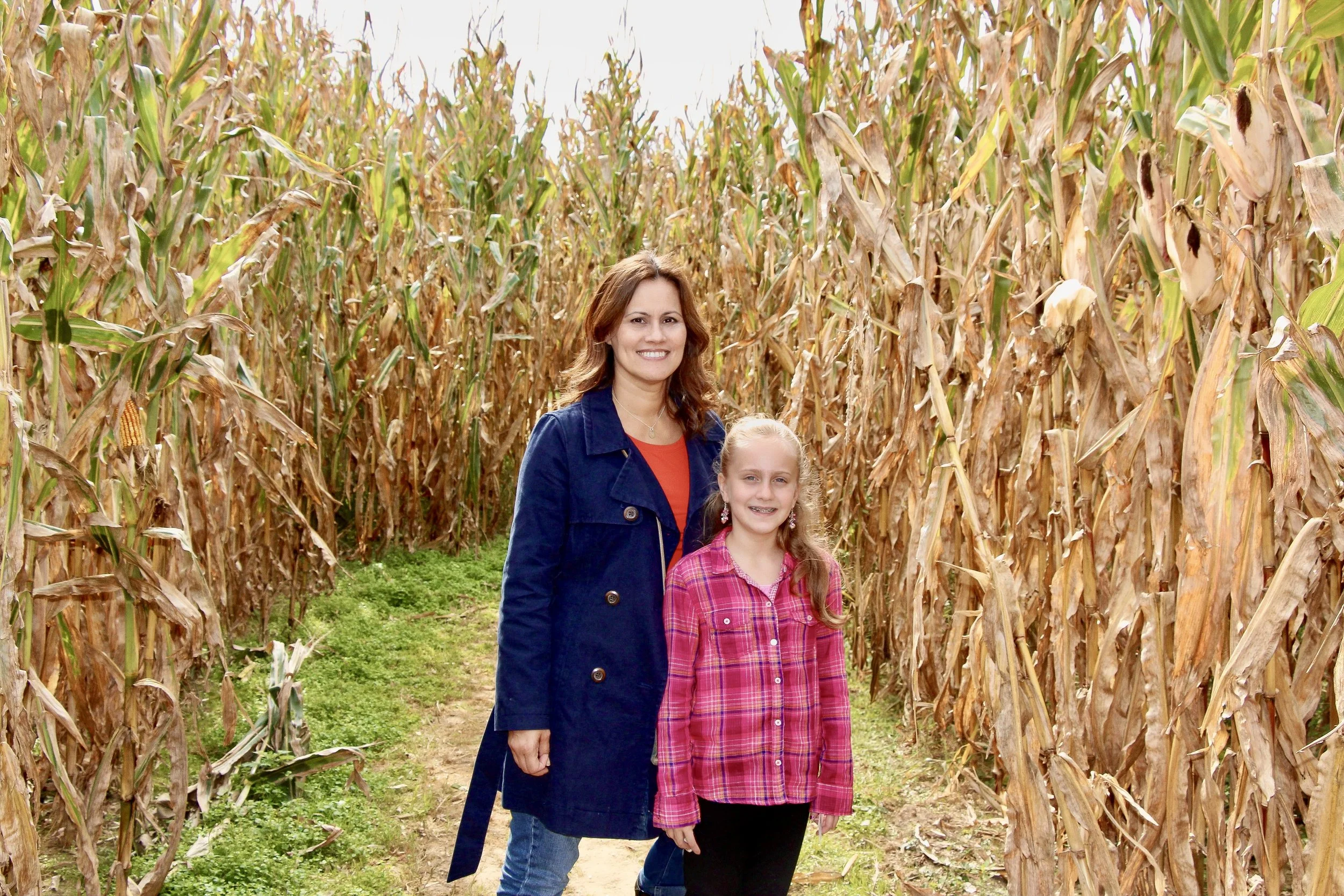 Woman in a navy coat and girl in a pink plaid shirt smiling together on a path through a corn maze lined with golden cornstalks