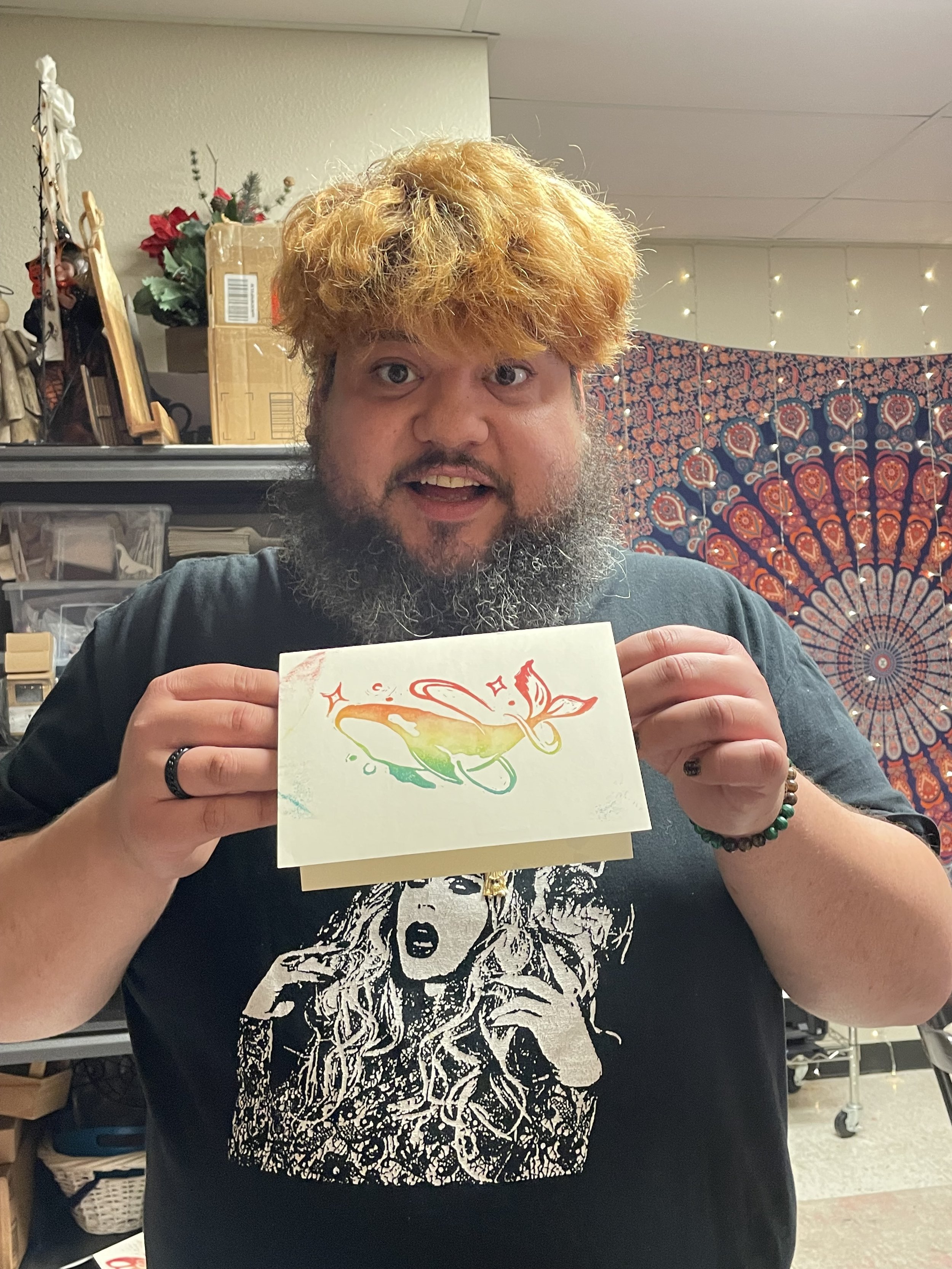 Man with rainbow-colored hair and a beard holding a greeting card with a rainbow-colored butterfly design, standing in a room decorated with string lights and a vibrant tapestry.