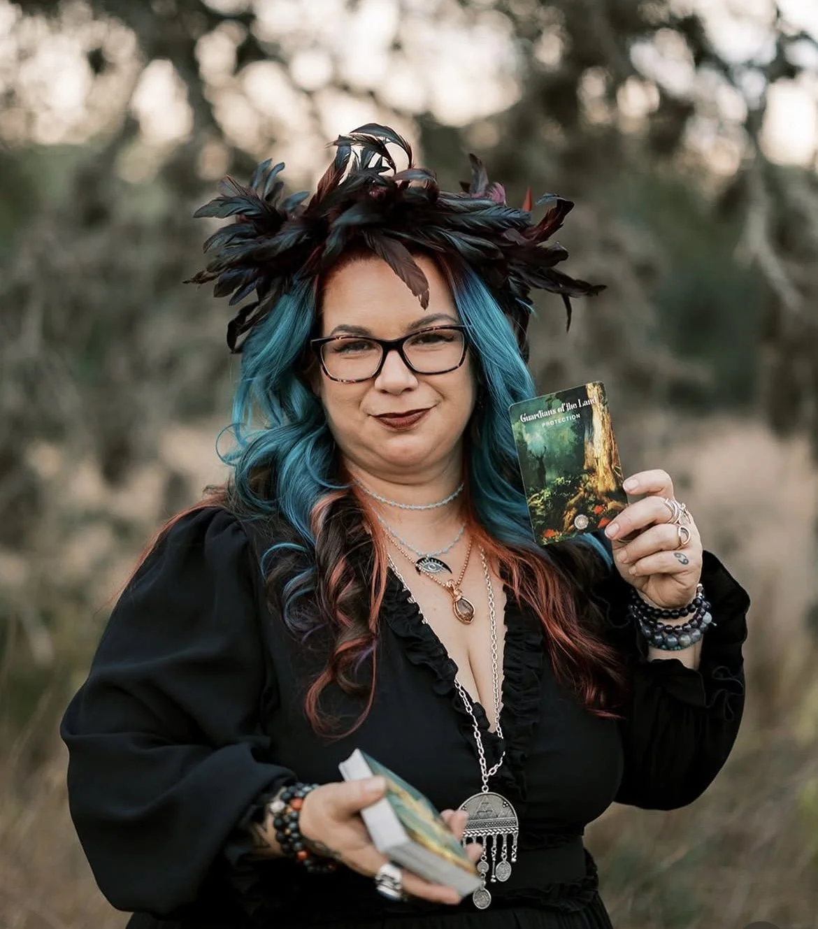 A woman with blue and red hair, glasses, and dark lipstick, wearing a black dress and adorned with various necklaces and bracelets, holds tarot cards in an outdoor setting with blurred natural background. She wears a large feathered hat with dark feathers.