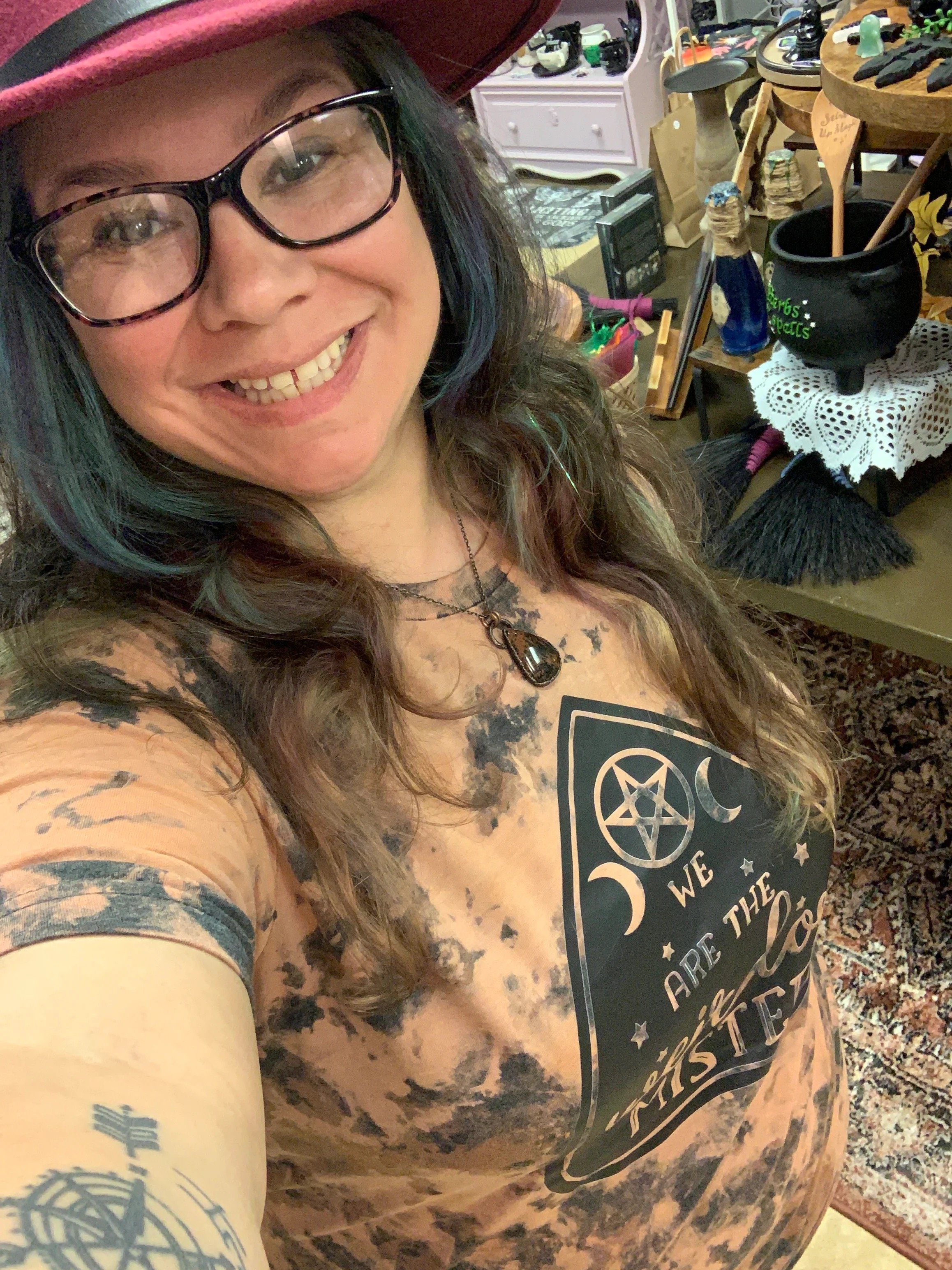 A woman with glasses and a red hat taking a selfie in a room filled with various items, including a table with a black pot, a doily, and decorative objects.