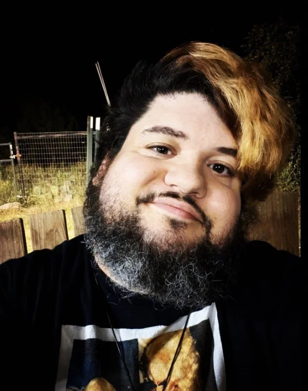 A man with a beard and styled hair, smiling at the camera, outdoors at night.
