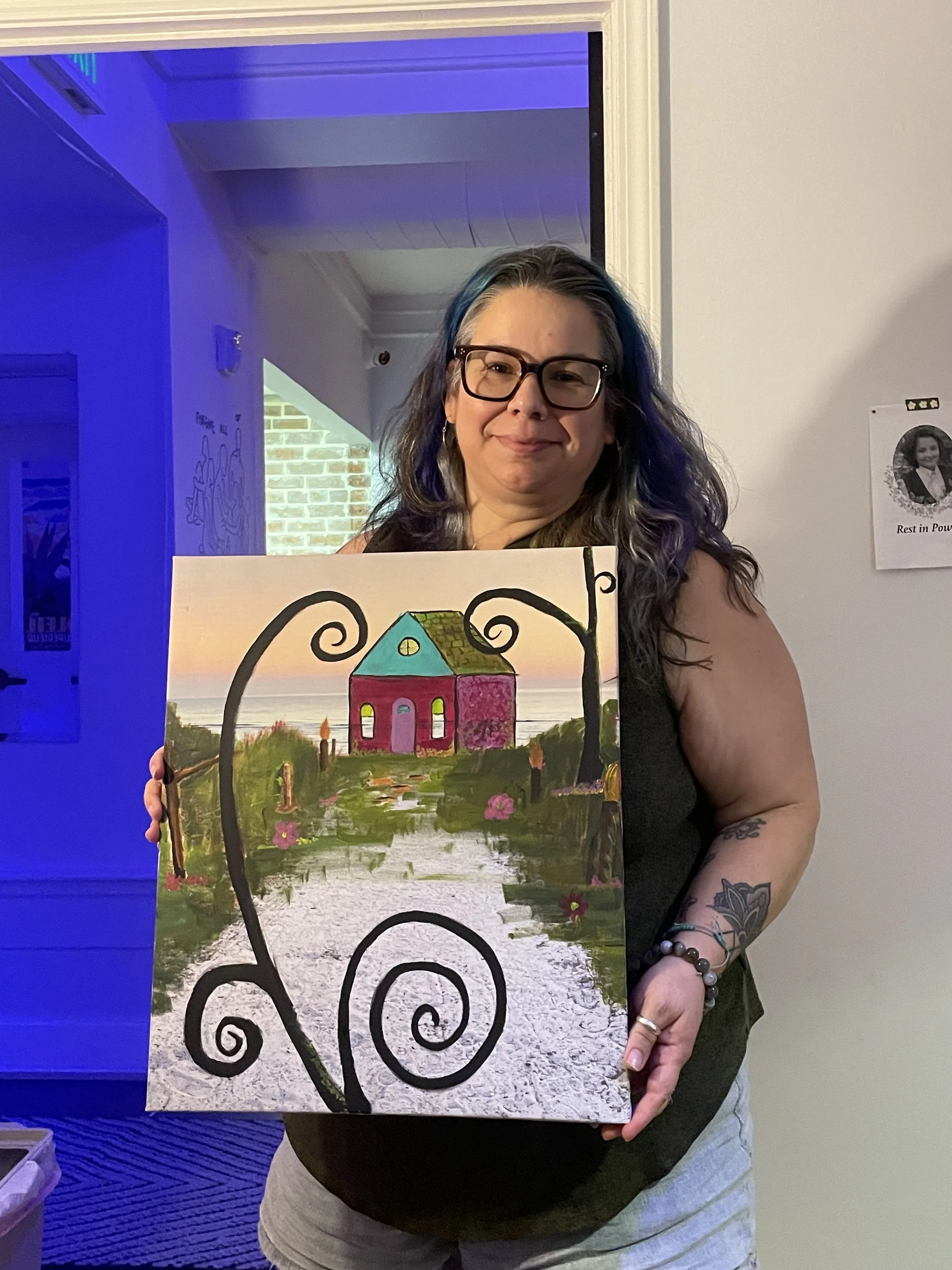 Woman with glasses and blue-highlighted hair holding a colorful painting of a pink house with a green roof, trees, and a pathway, against a sunset background.