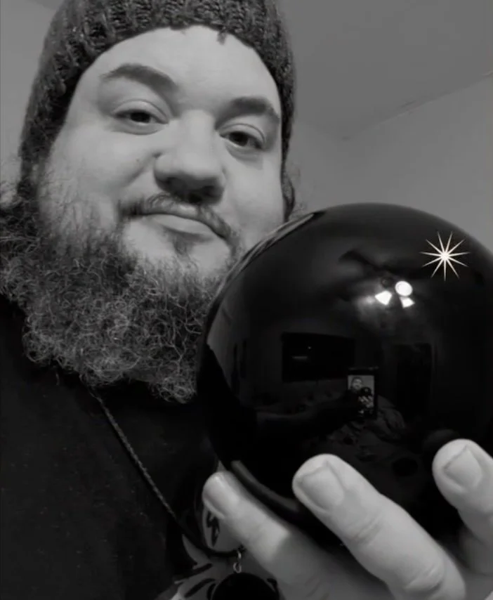 A man with a beard and wearing a knit cap taking a photo of himself with a shiny black spherical object, possibly a bowl or decorative item, in a room with a reflected ceiling light visible in the sphere.
