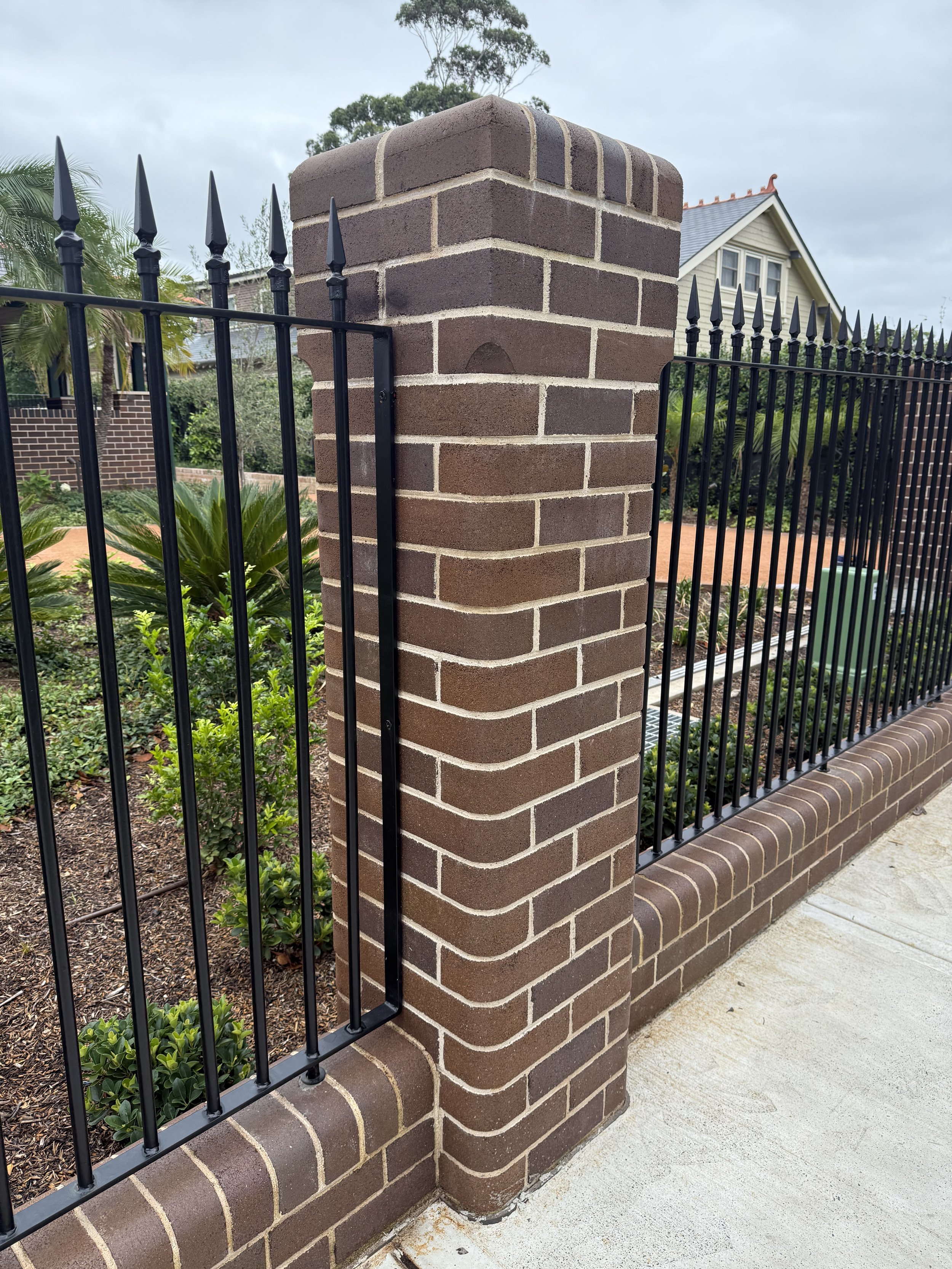Close-up of a brick fence post with decorative black metal fencing and a sidewalk in the foreground.