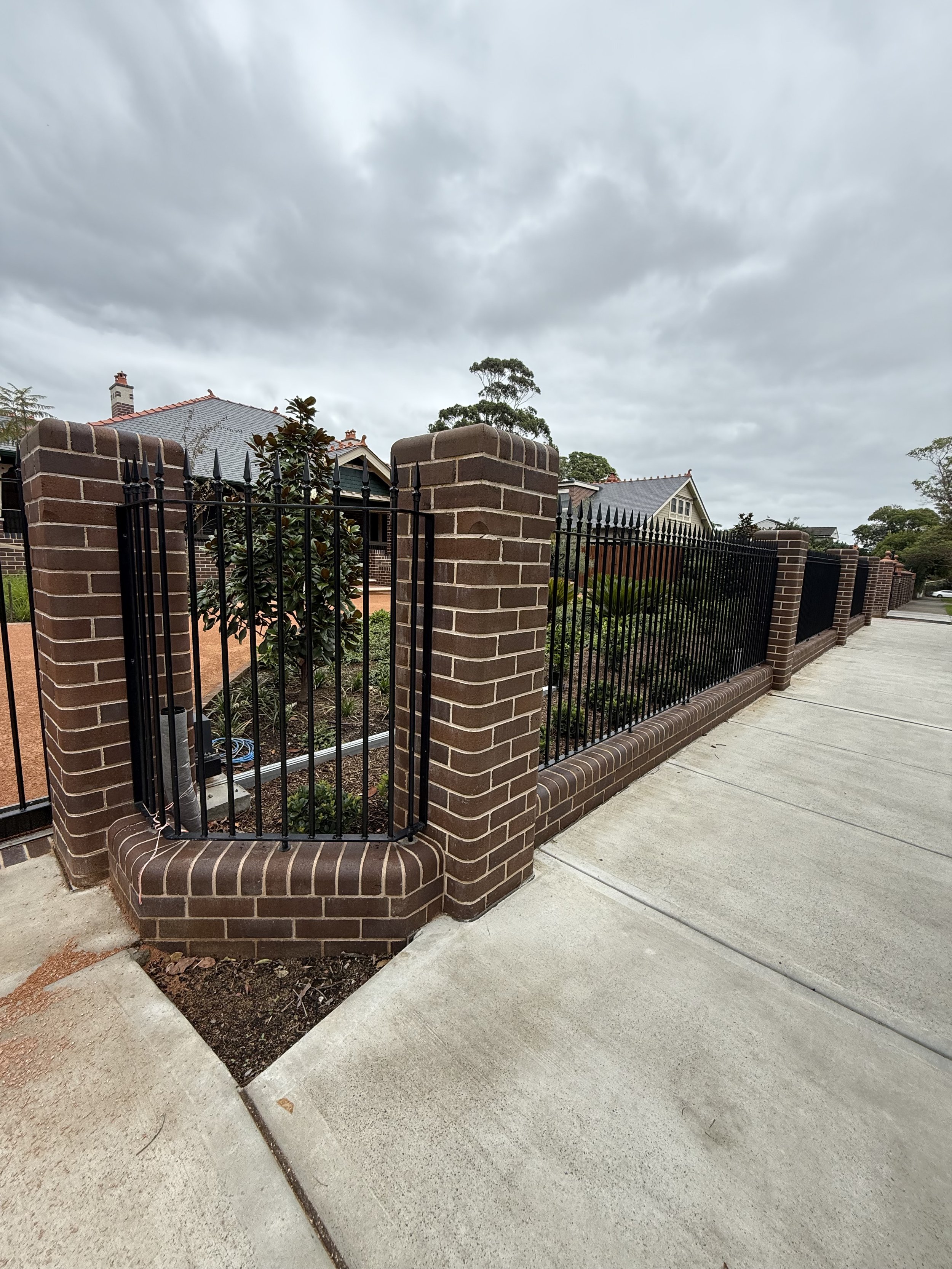 View of a brick fence with black metal fencing and a small gate, surrounding a garden with plants and a house with a grey roof in the background, under an overcast sky.