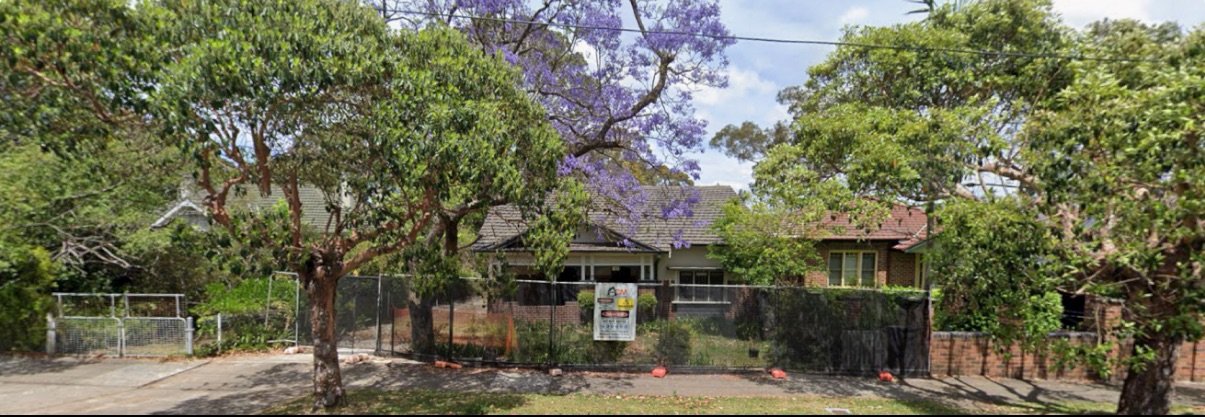 A house under construction or renovation, surrounded by a black fence, with trees and greenery in the front yard.