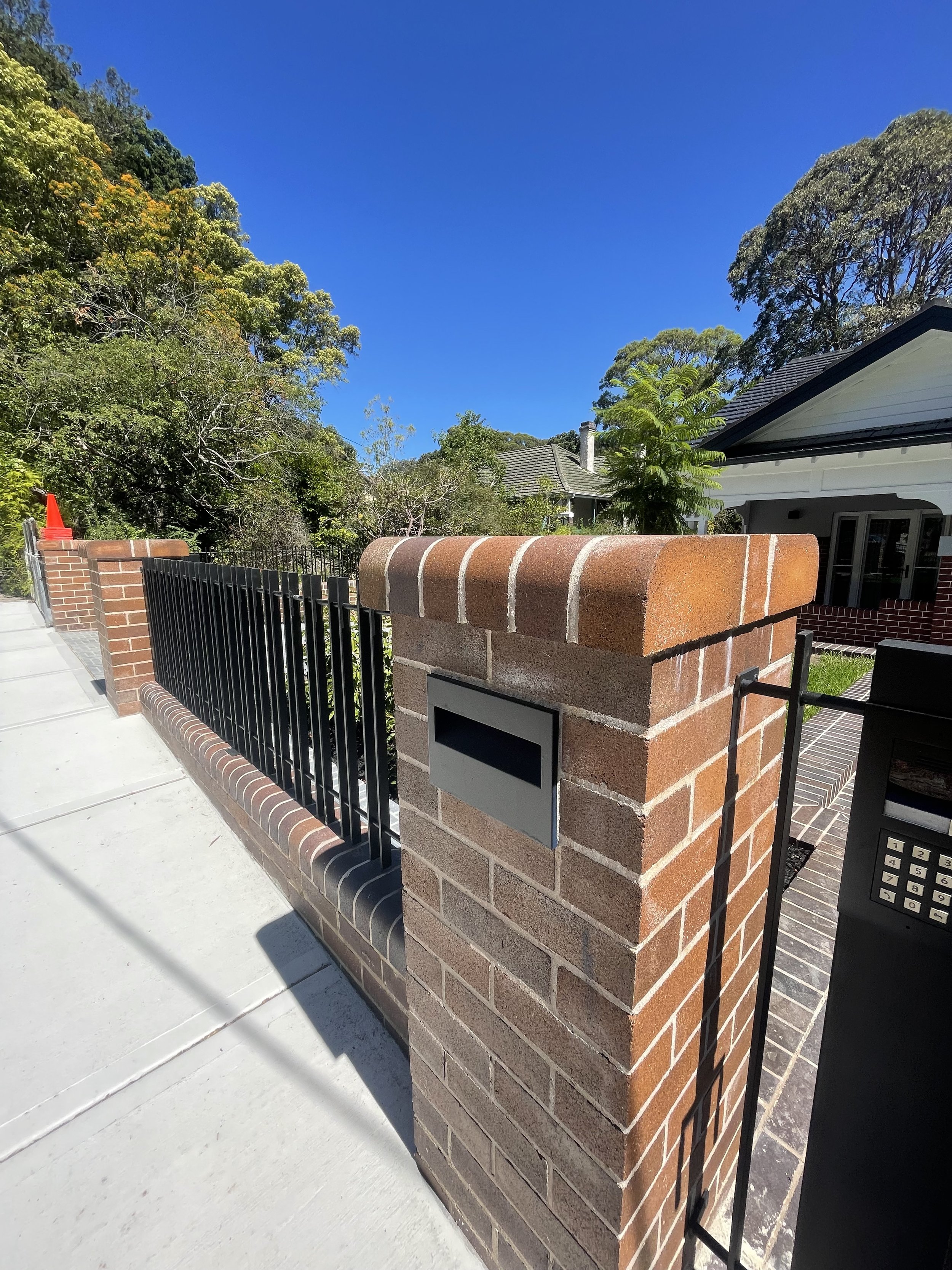 Close-up of a brick gate pillar with a mailbox and black metal fencing, yellow and green trees in the background under a clear blue sky.