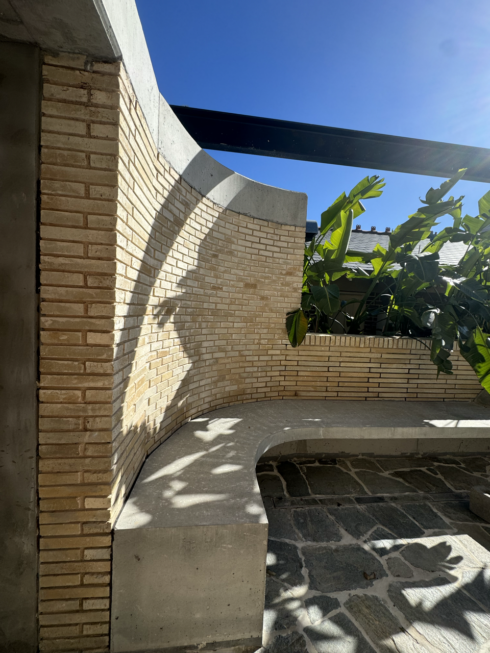 Outdoor brick and concrete curved bench with shadows, green leafy plants, blue sky, modern building elements.