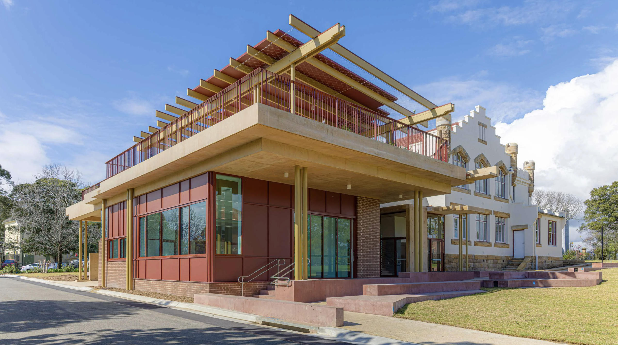 Modern multi-story building with large glass windows, red accents, and a prominent balcony with a metal railing, set against a bright blue sky.