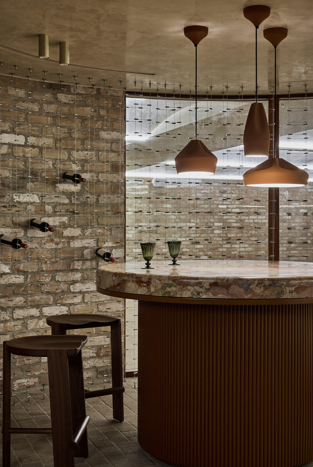 Modern interior with a brick wall, a marble-topped bar, hanging pendant lights, wine bottles on a vertical wine rack, and two green glasses on the bar counter.