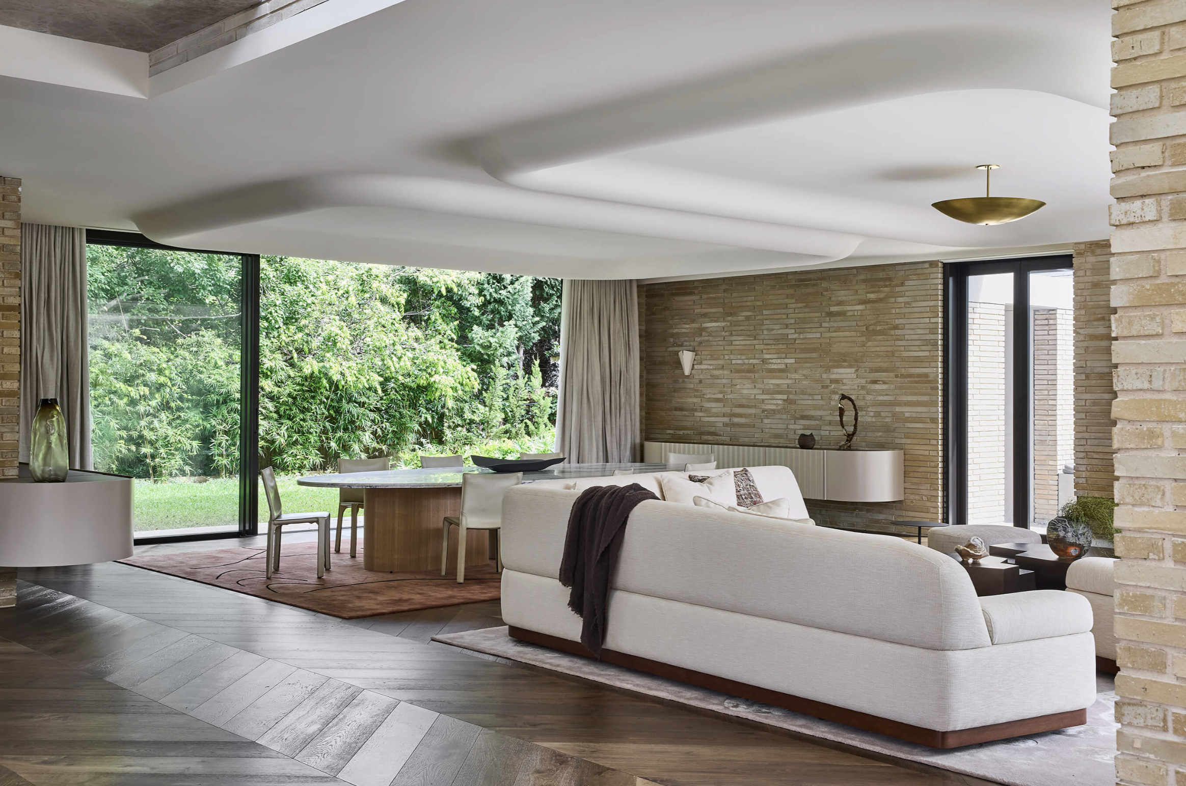 Modern living room with a white sofa, a round wooden dining table, and large windows revealing greenery outside.