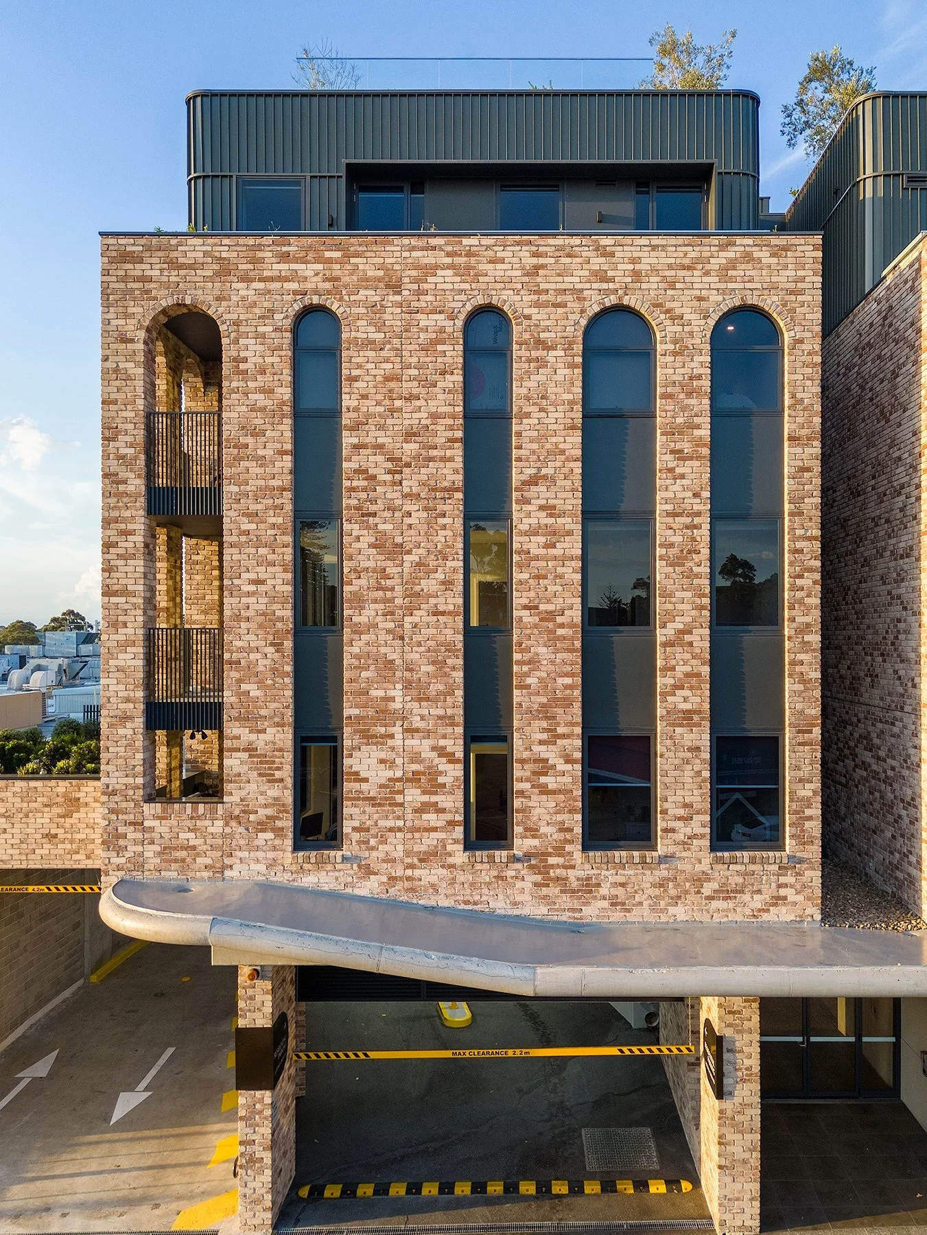 Modern brick building with tall, narrow arched windows and balconies, parking area with clearance bar, and a curved concrete driveway.