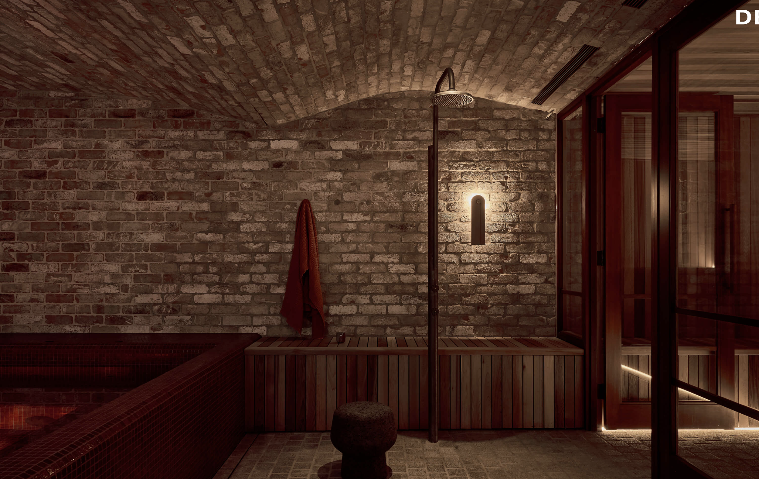 Indoor shower area with brick arched ceiling, including a rain shower head, a red towel hanging, a small stool, and a corner with red mosaic tiling.