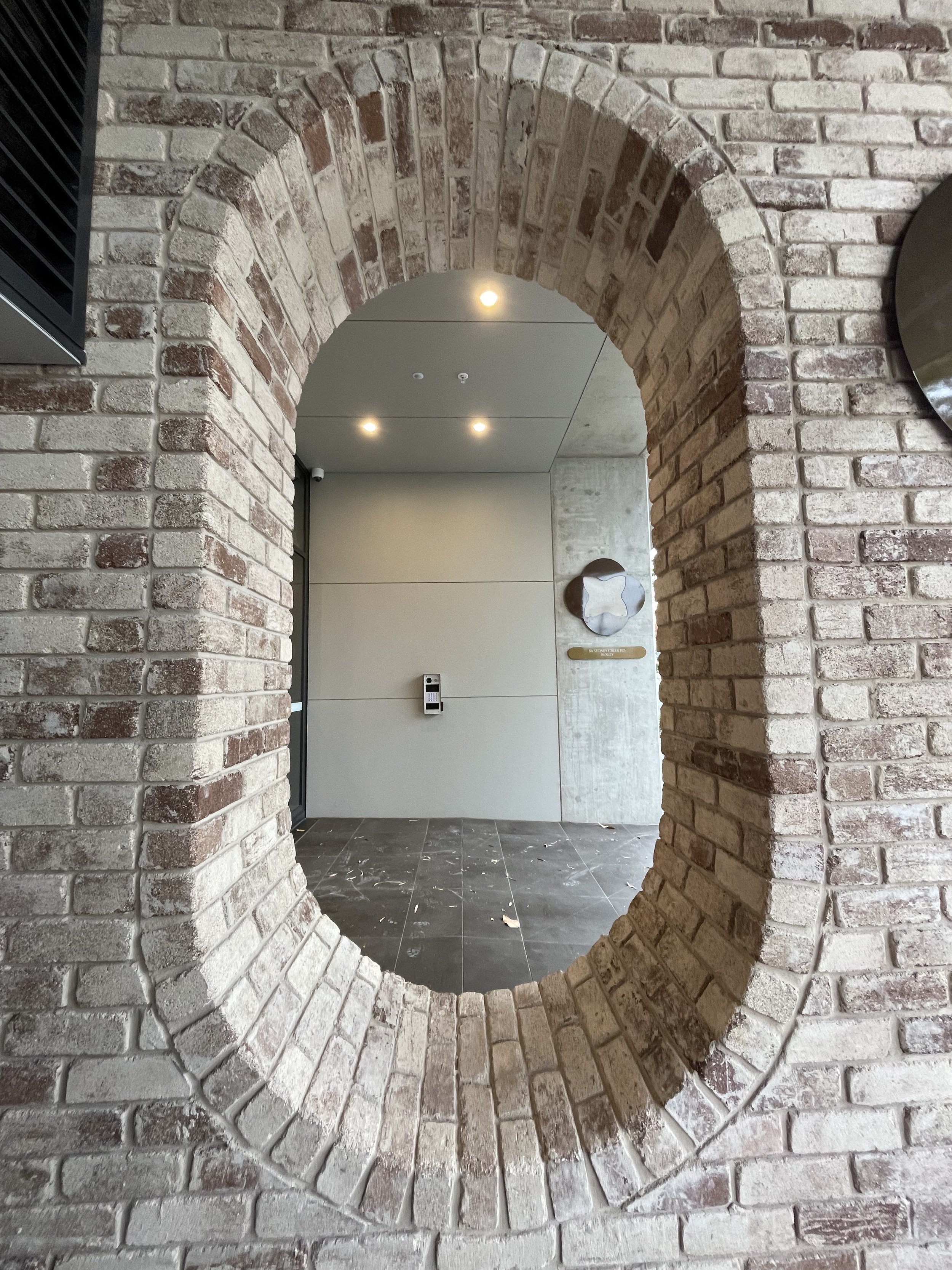 A brick archway with an oval opening, framing a modern interior space with a light-colored wall, a security keypad, and a metallic logo on the wall.