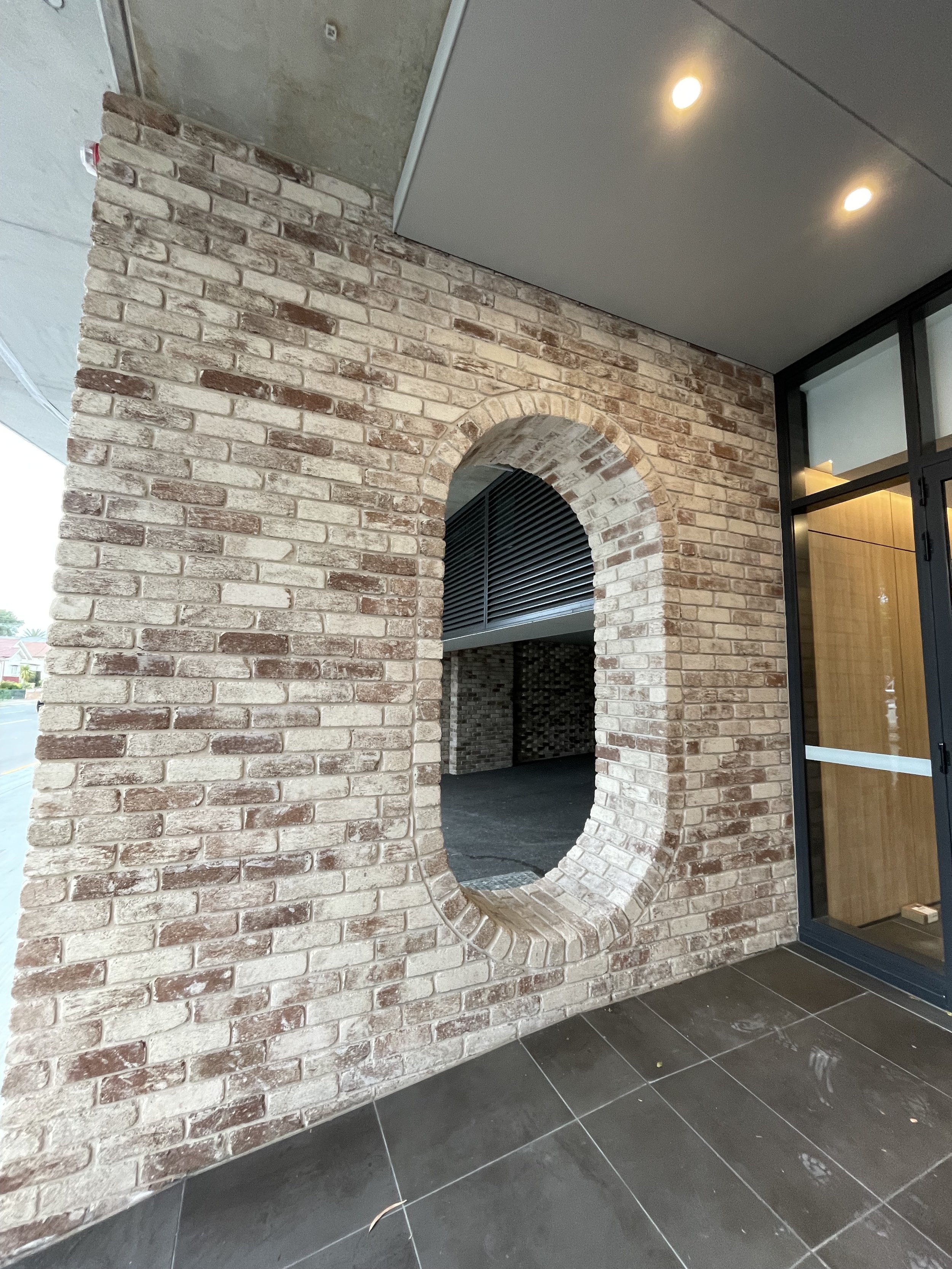 A brick wall with an oval-shaped opening, part of a modern interior with dark tiled flooring and ceiling lighting.