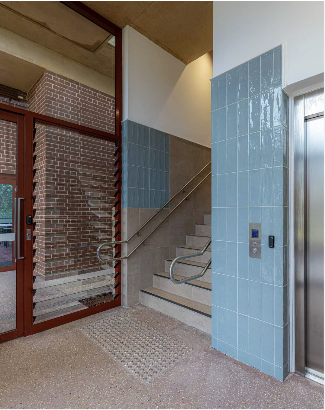 Interior of a building lobby with stairs, blue tiled wall, brick wall, an elevator, and a glass door.