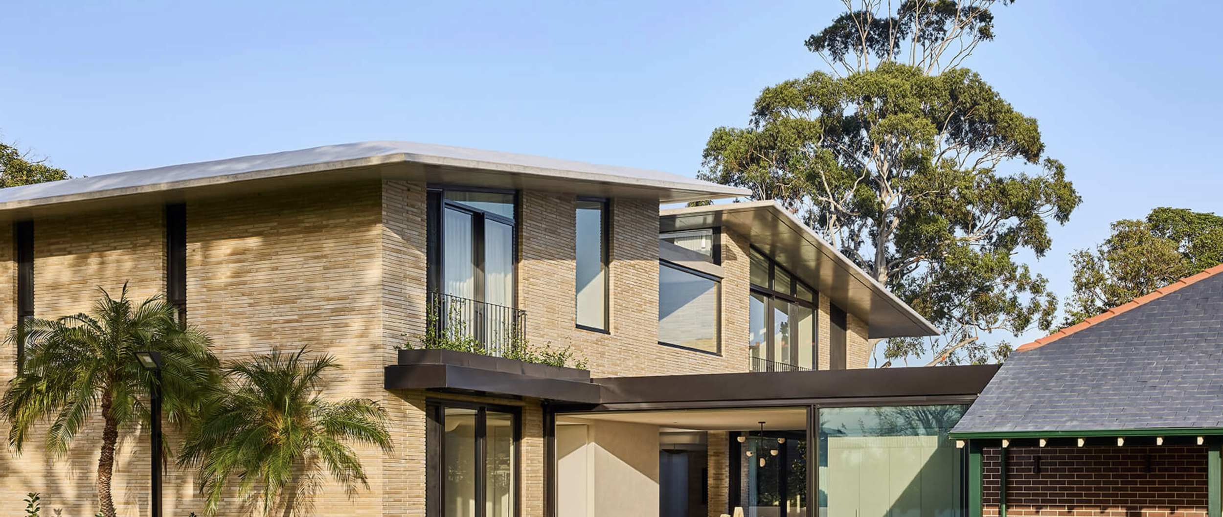 Modern two-story house with large windows and brick exterior, surrounded by trees and palm trees under a clear blue sky.