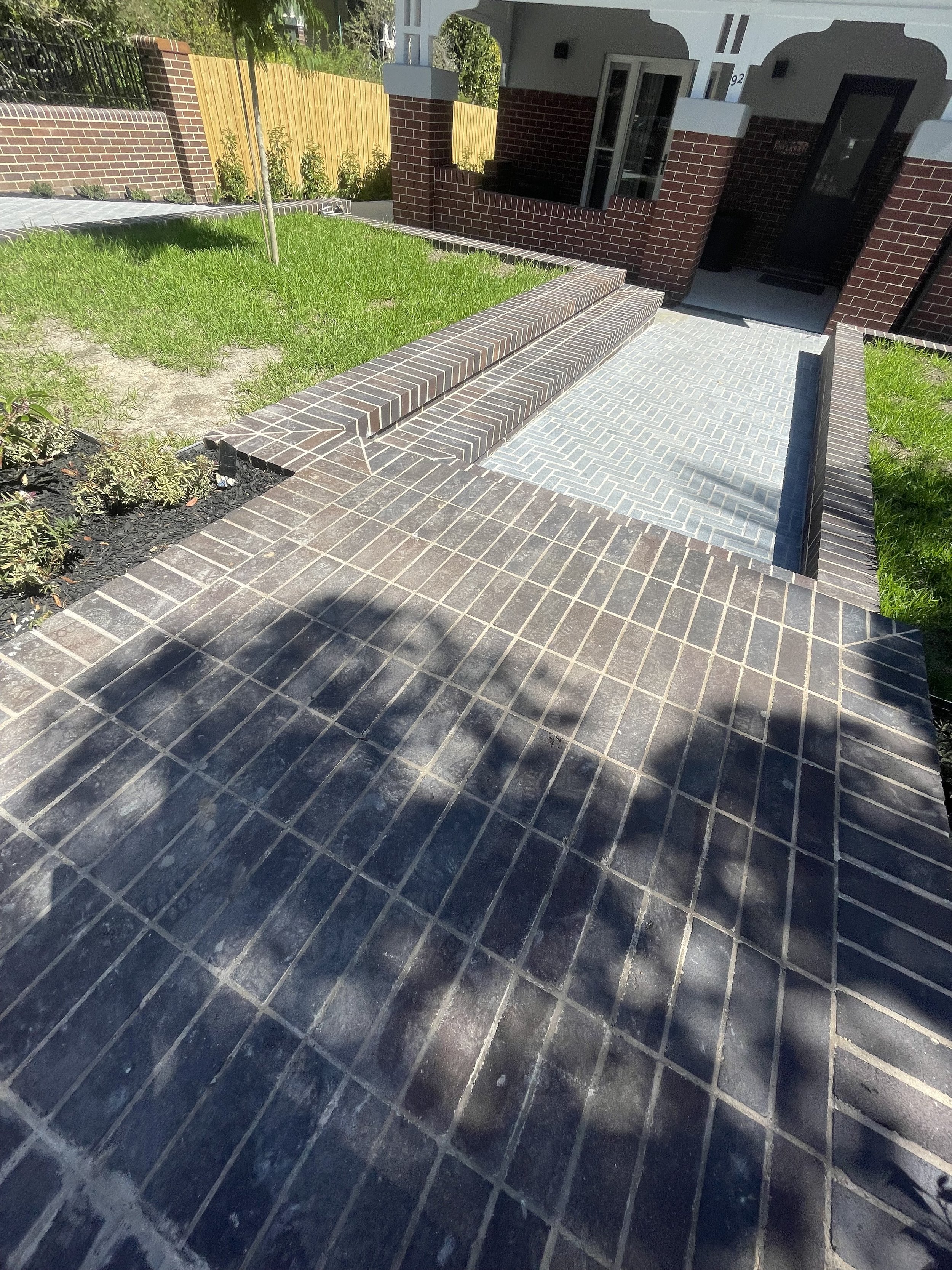 View of the entrance to a house with a brick walkway, steps, and porch, shaded by a tree casting shadow, with grass and a wooden fence in the background.