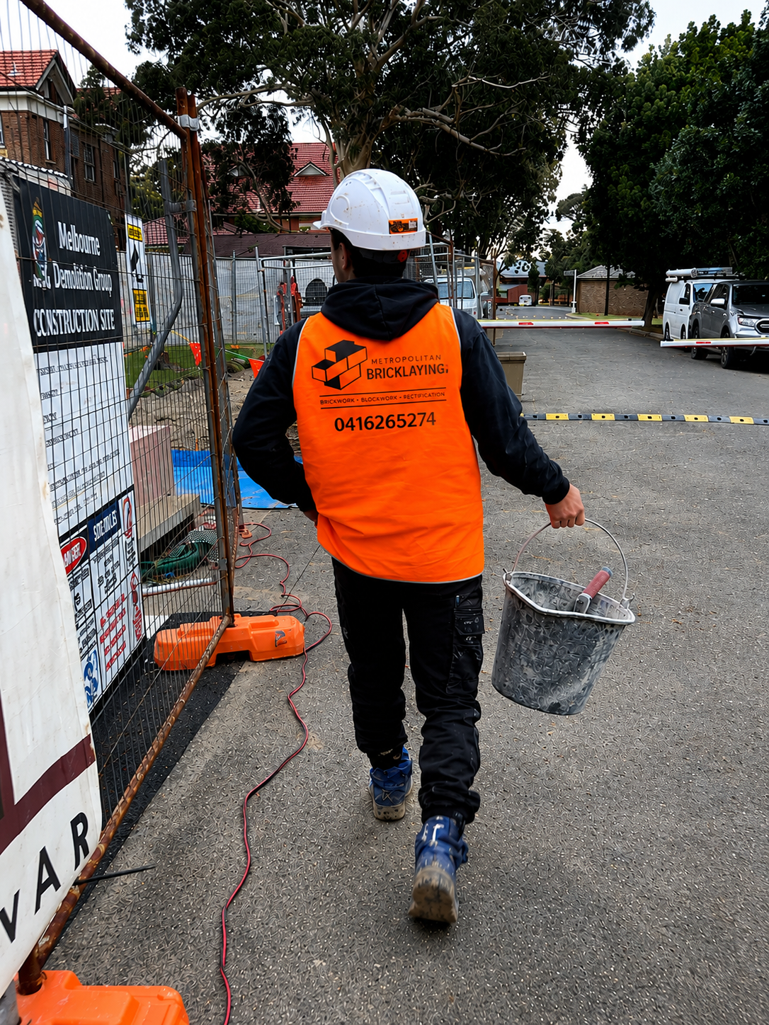 A construction worker in an orange vest and white helmet walking away from the camera, carrying a bucket with a paint roller inside on a construction site with fencing and parked cars.