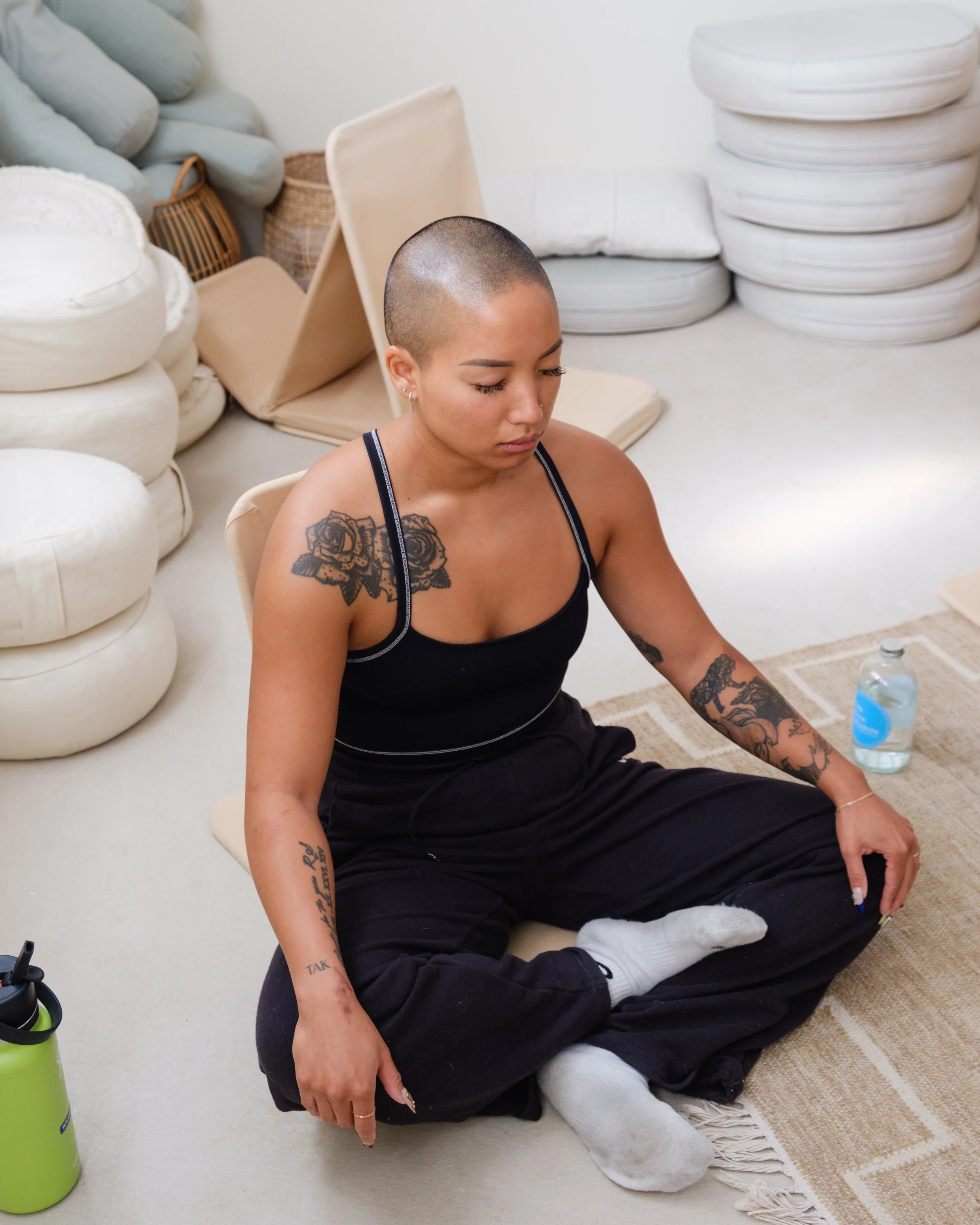 Person meditating indoors, sitting cross-legged in a minimalist room with cushions and bottled water nearby.