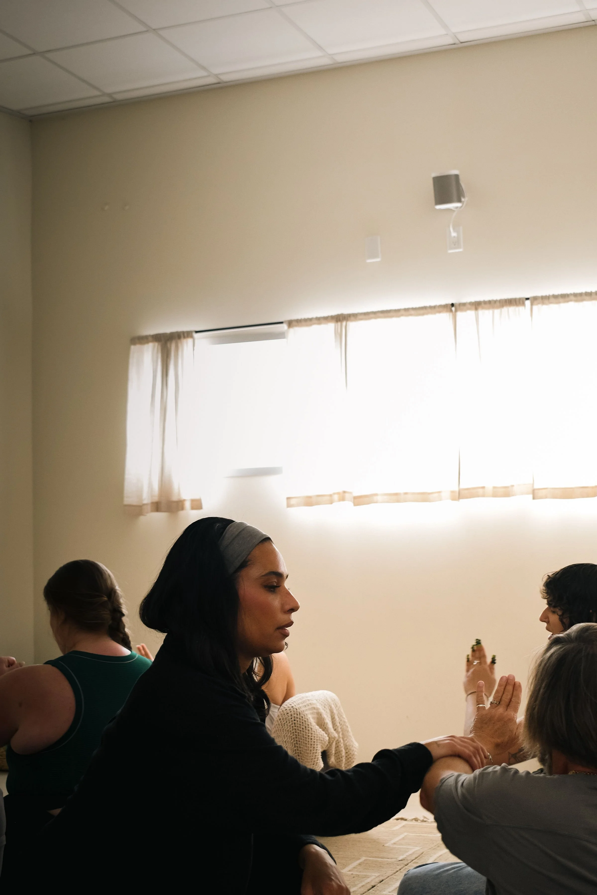 A woman with dark hair and a gray headband sitting on the floor, with her eyes closed, holding hands with an older woman, in a room with beige walls, a white ceiling, and a window with light-colored curtains.