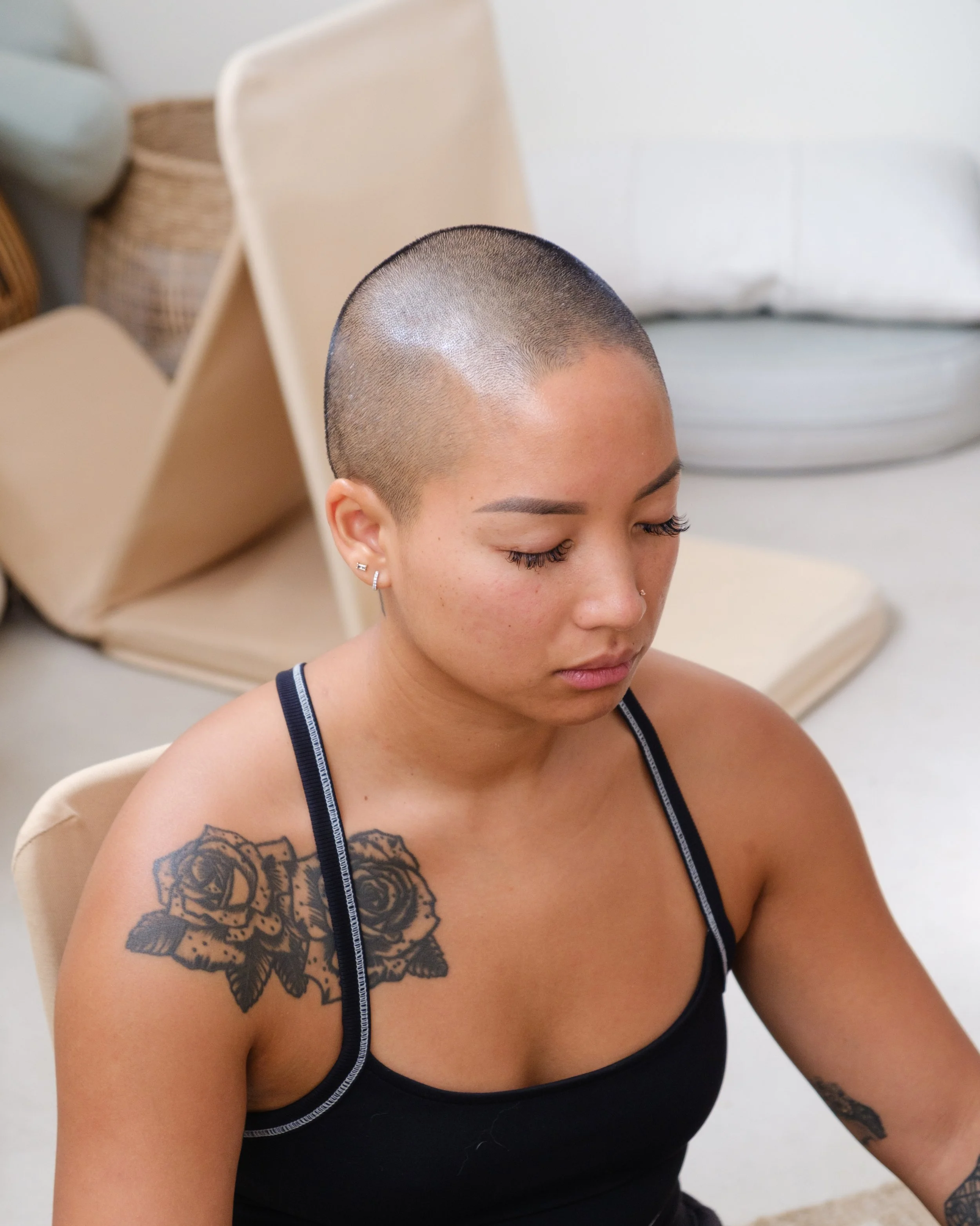 A woman with a shaved head, tattoos on her shoulder and arm, wearing earrings and a black tank top, sitting in a room with beige chairs and a wicker basket in the background.