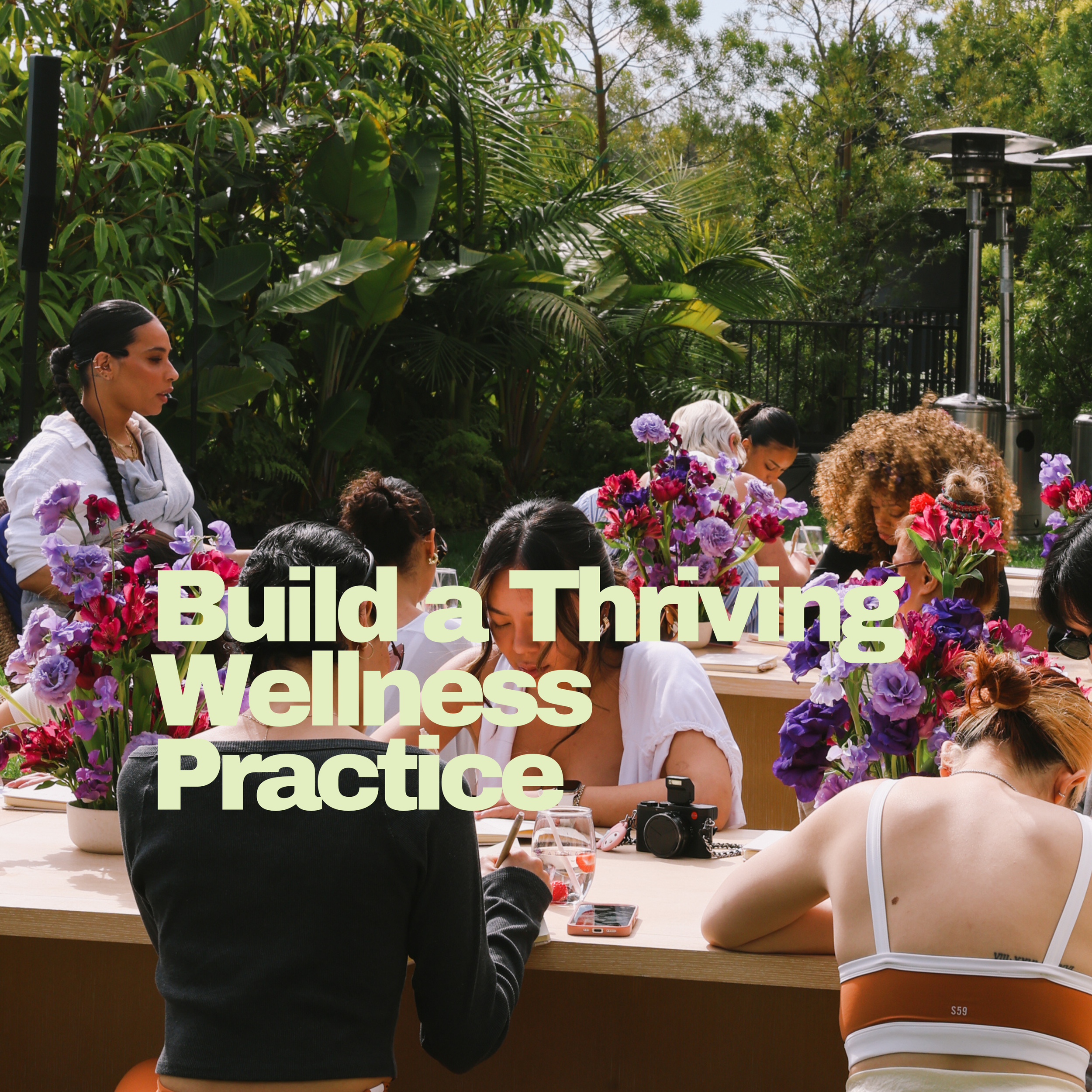 Group of people sitting at a table outdoors surrounded by lush greenery and colorful flowers, engaging in a wellness practice or activity.