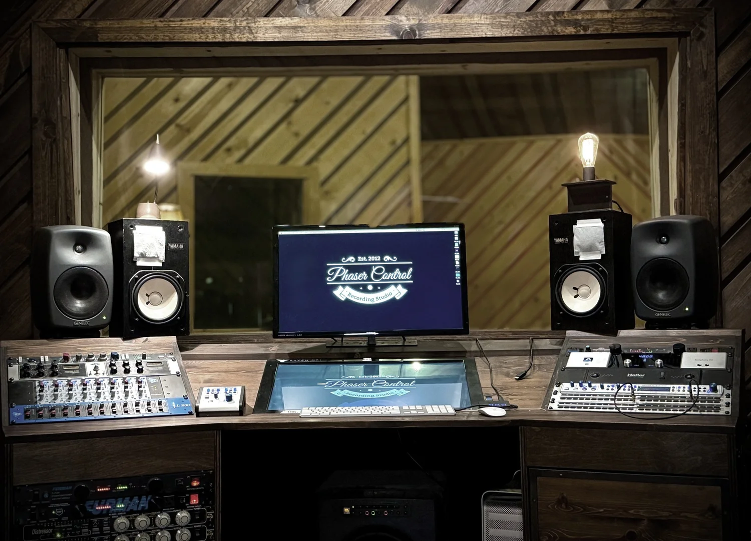 The Phaser Control desk — mixing console and monitors under warm light, with the live room visible through the control-room window behind.