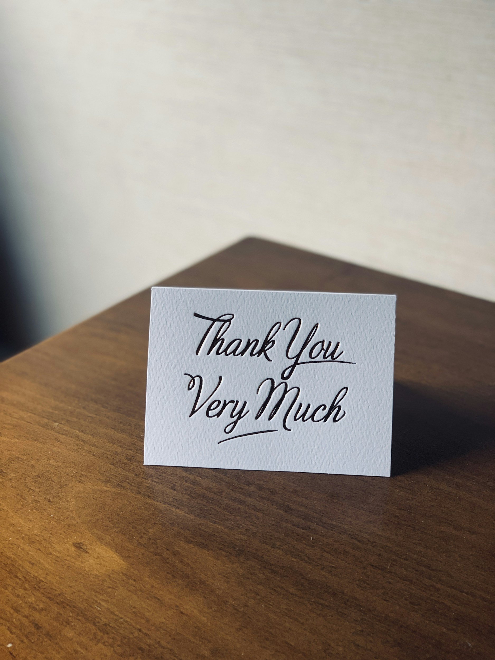 A "Thank You Very Much" card placed on a wooden table, featuring elegant black cursive writing on a white background.