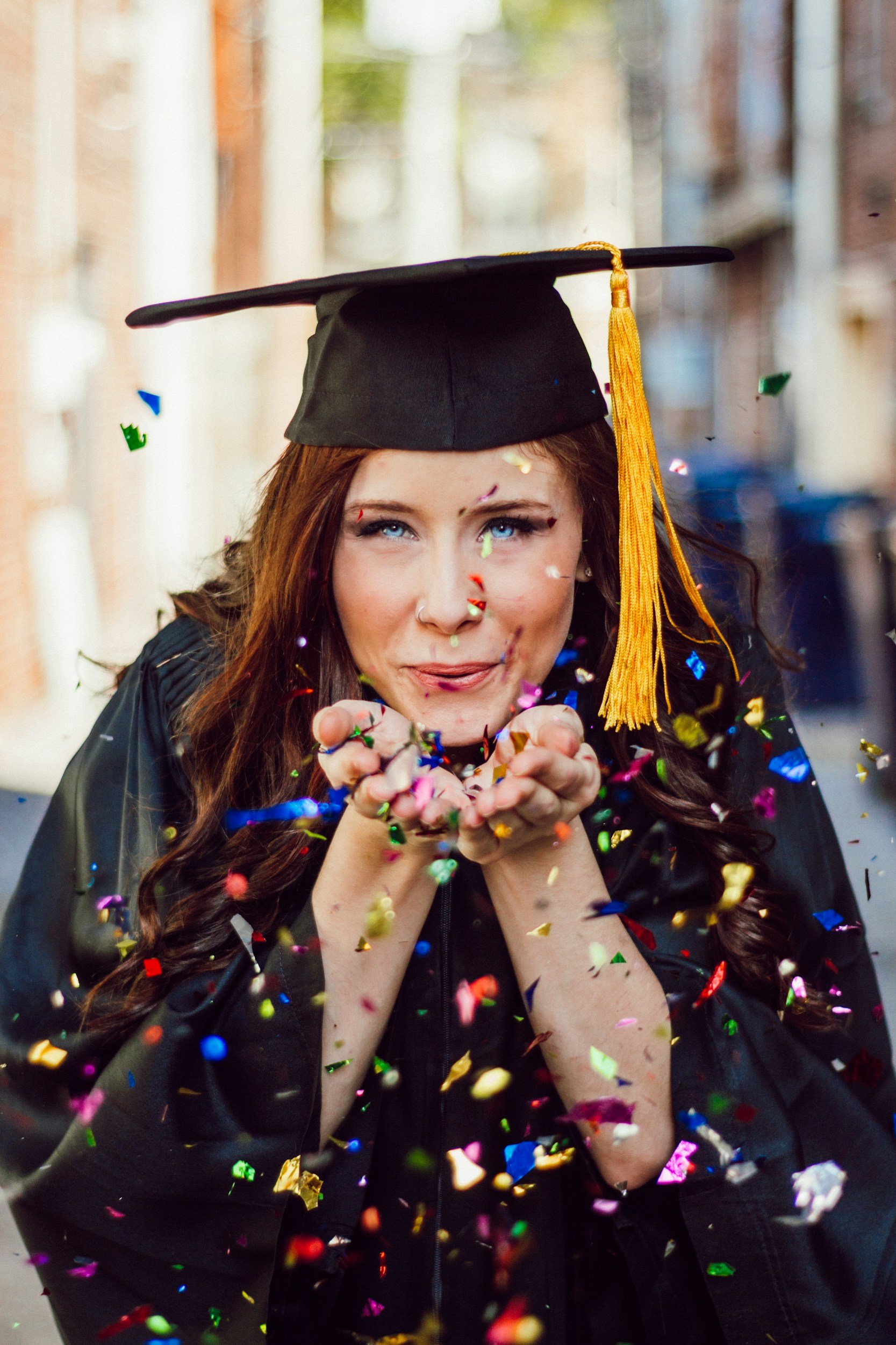 A graduate woman in a cap and gown blowing colorful confetti.