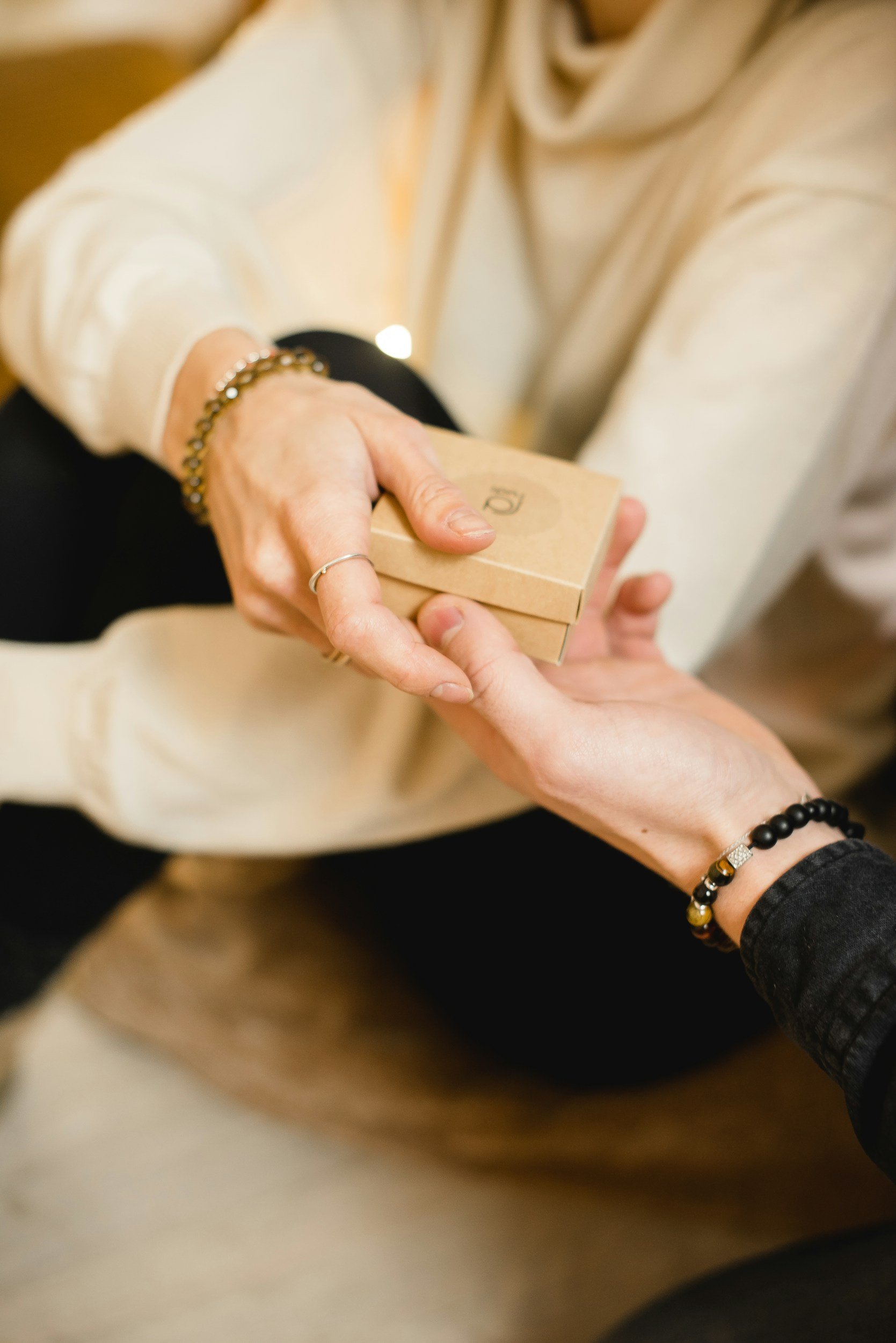 Two people exchanging a small, brown box, sitting on a couch. One has a bracelet and rings on.