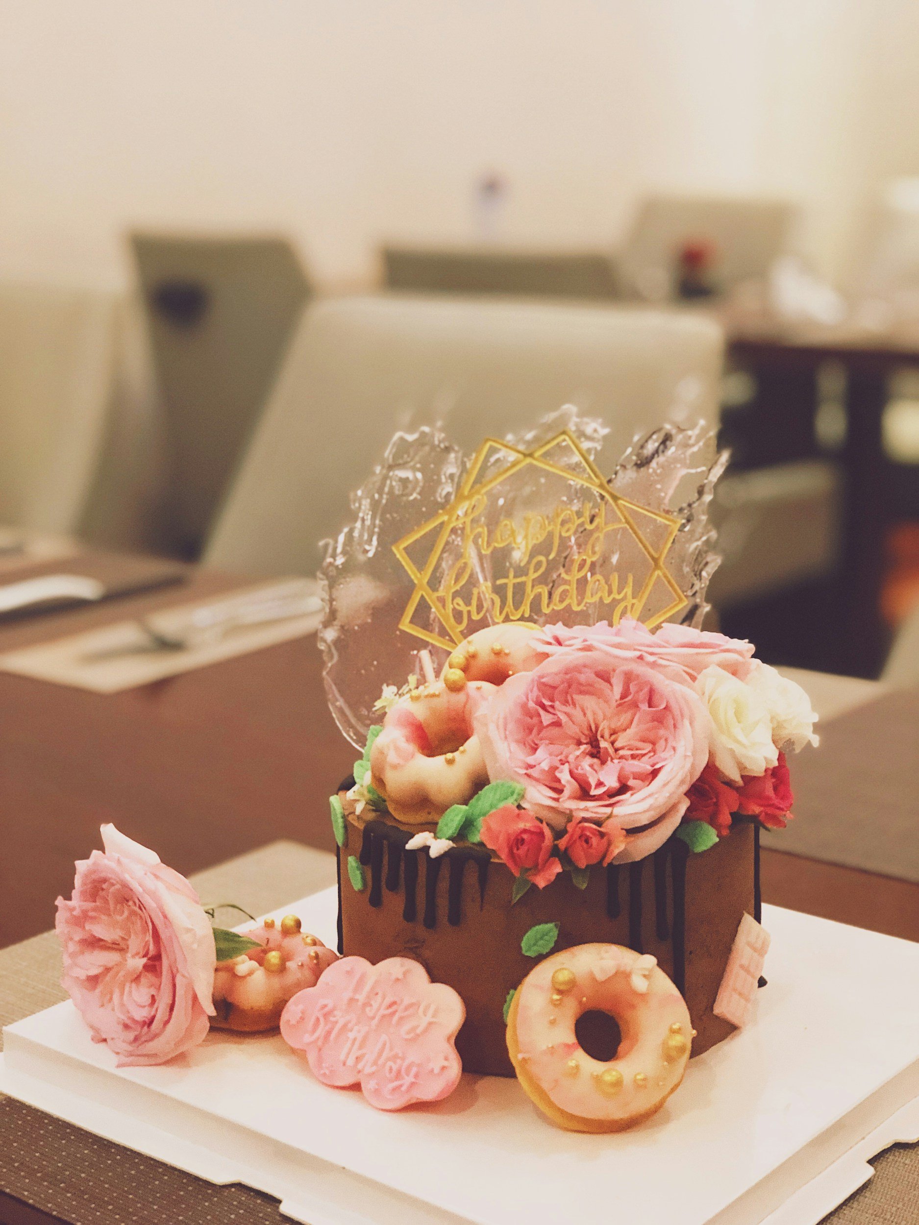 Decorated birthday cake with pink flowers and donuts on top, chocolate drip design, and a "Happy Birthday" topper.