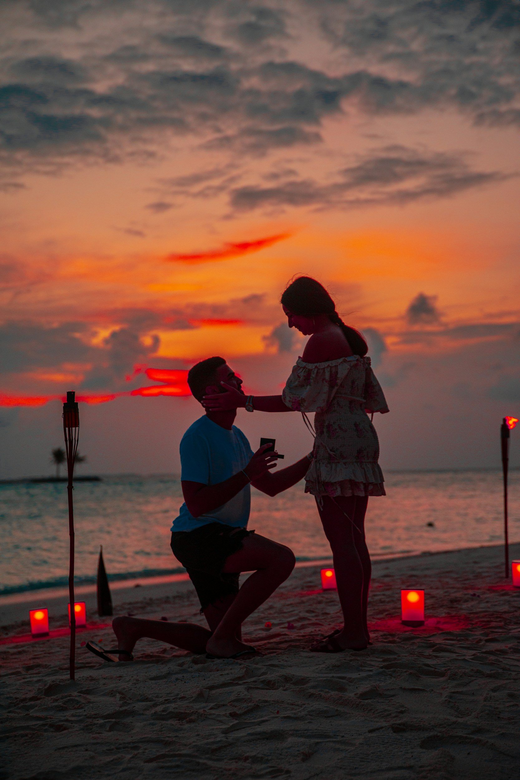 A romantic beach proposal at sunset with a man kneeling and holding a ring, surrounded by candles and tiki torches.