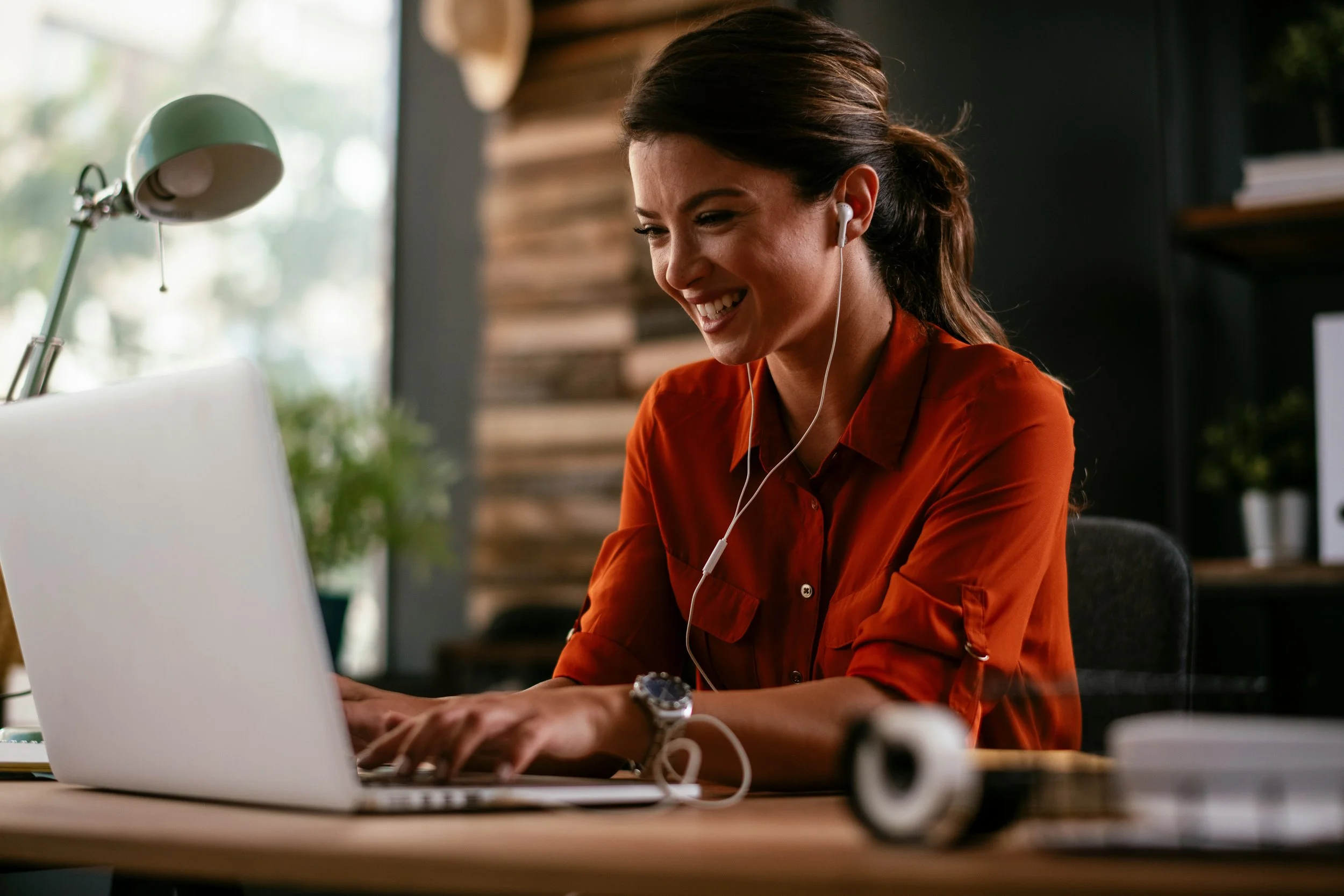 Mujer sonriendo mientras usa audífonos y trabaja en una laptop en un ambiente moderno con iluminación natural.