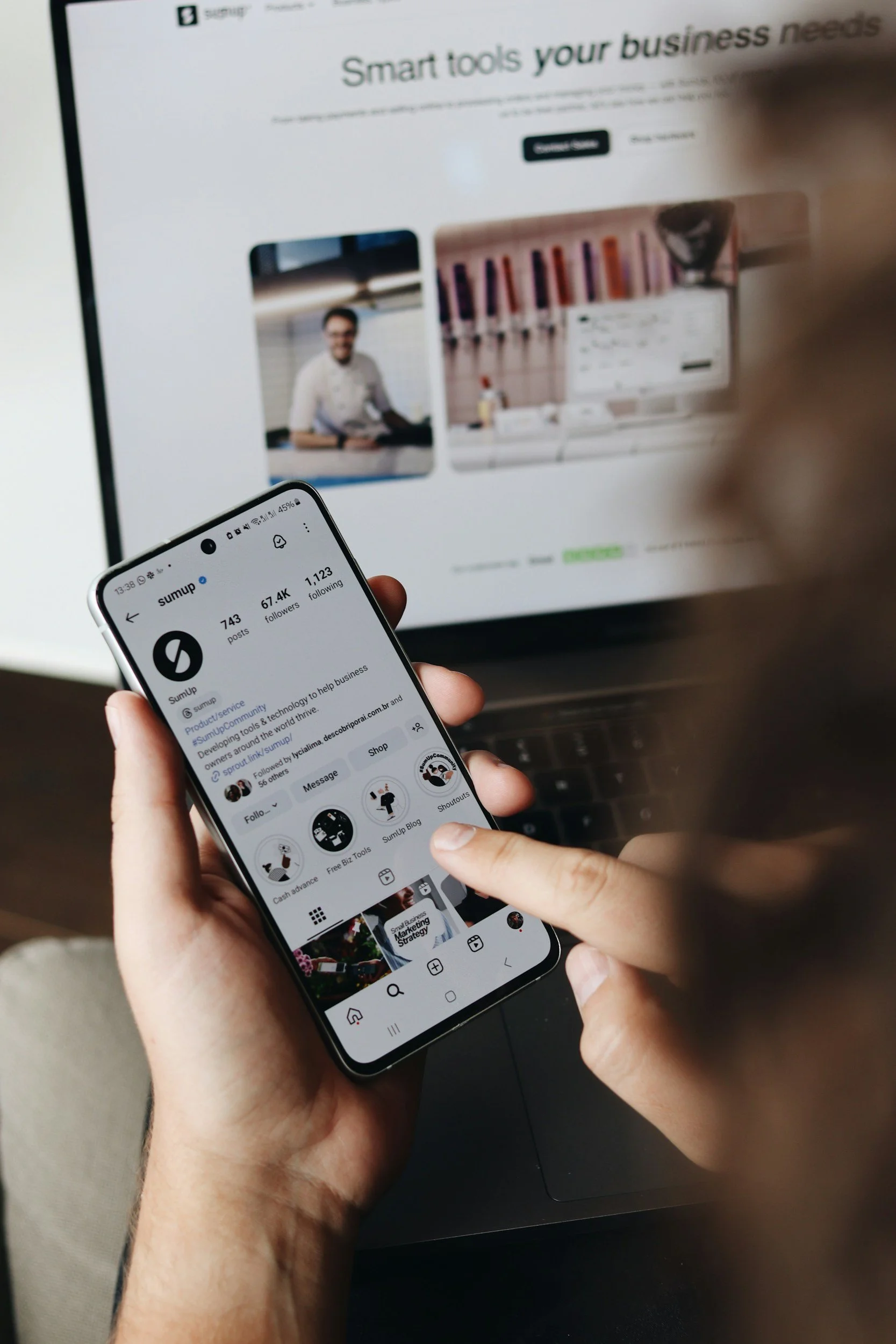Persona sosteniendo un teléfono móvil que muestra la página de perfil de la red social 'Sumup', con estadísticas de seguidores y publicaciones, en primer plano. En el fondo, una computadora con pantalla de contenido web borroso.