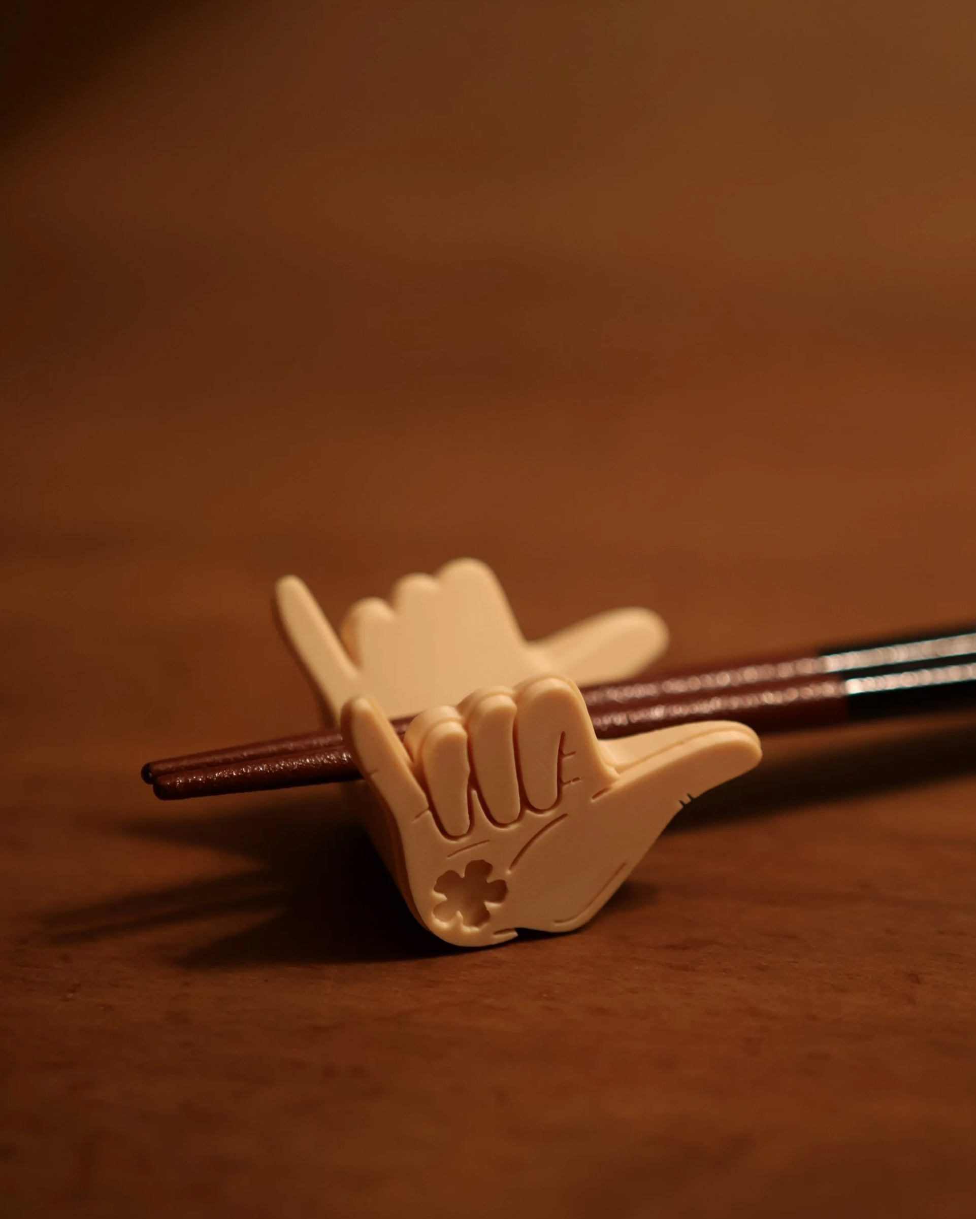 A chopstick holder (hashi) with a hand design, shaped like a shaka, holding a pair of plastic chopsticks, on a wooden surface.