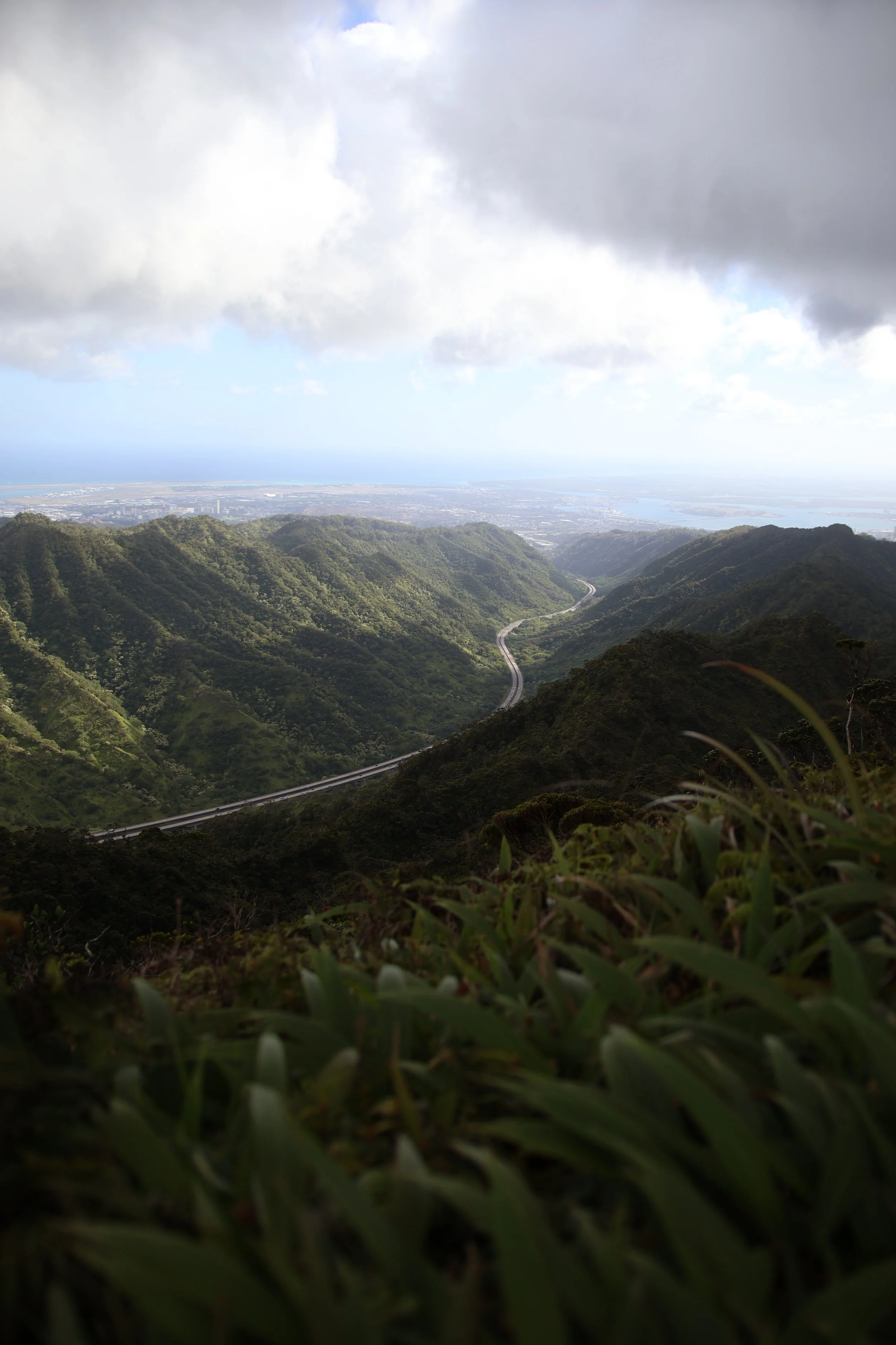 View of a lush, green mountain range with a winding road and distant cityscape under cloudy skies.