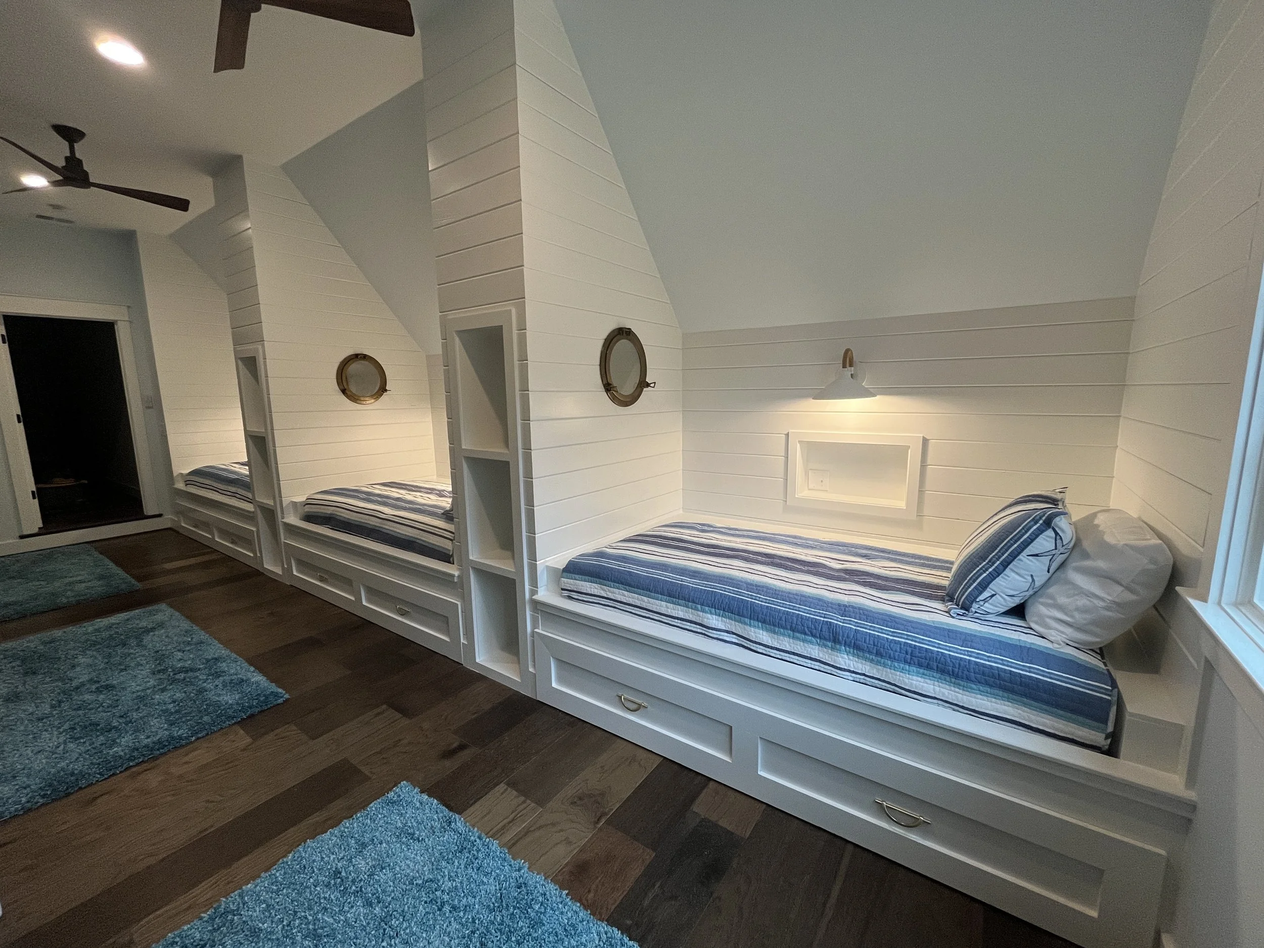 Cozy bedroom with built-in beds along a wall, each with striped bedding and pillows, wooden floors, and blue rugs, featuring wall-mounted lights and small decorative portholes.